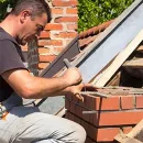 Worker laying bricks on a rooftop next to a chimney, using a trowel and level.