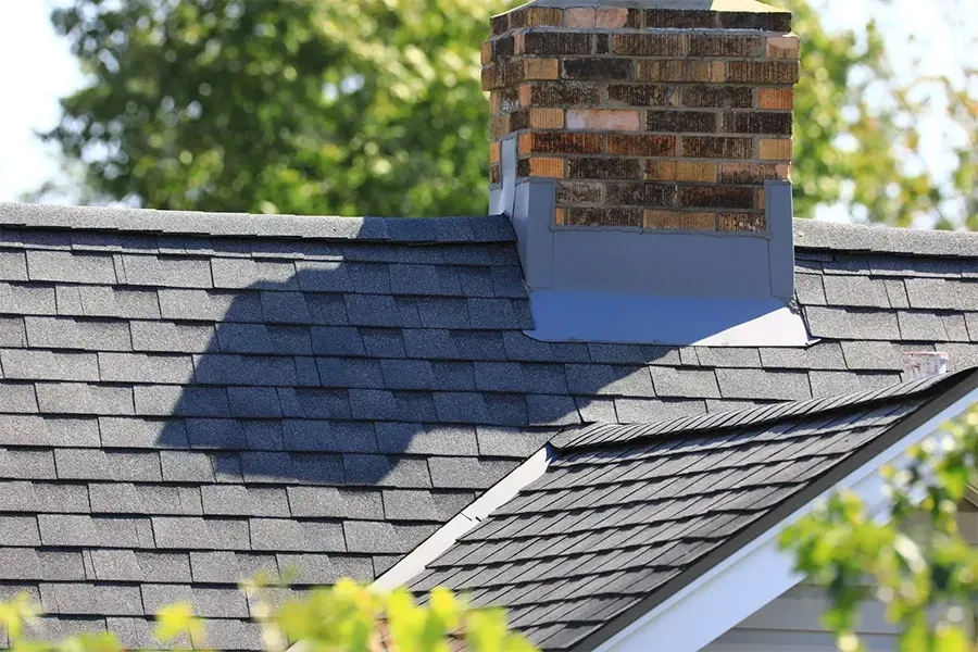 Roof with dark gray shingles and a brick chimney on a house, viewed through blurred green leaves.