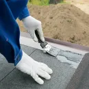 Gloved hands applying gray roofing sealant to a shingled roof seam