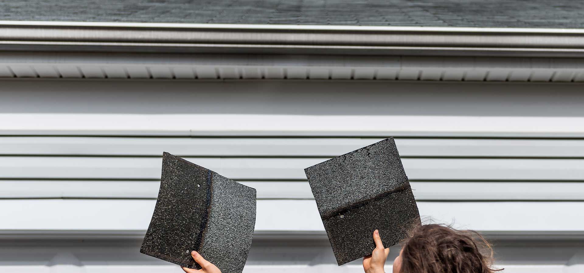 A woman is holding two pieces of shingles in front of a house.