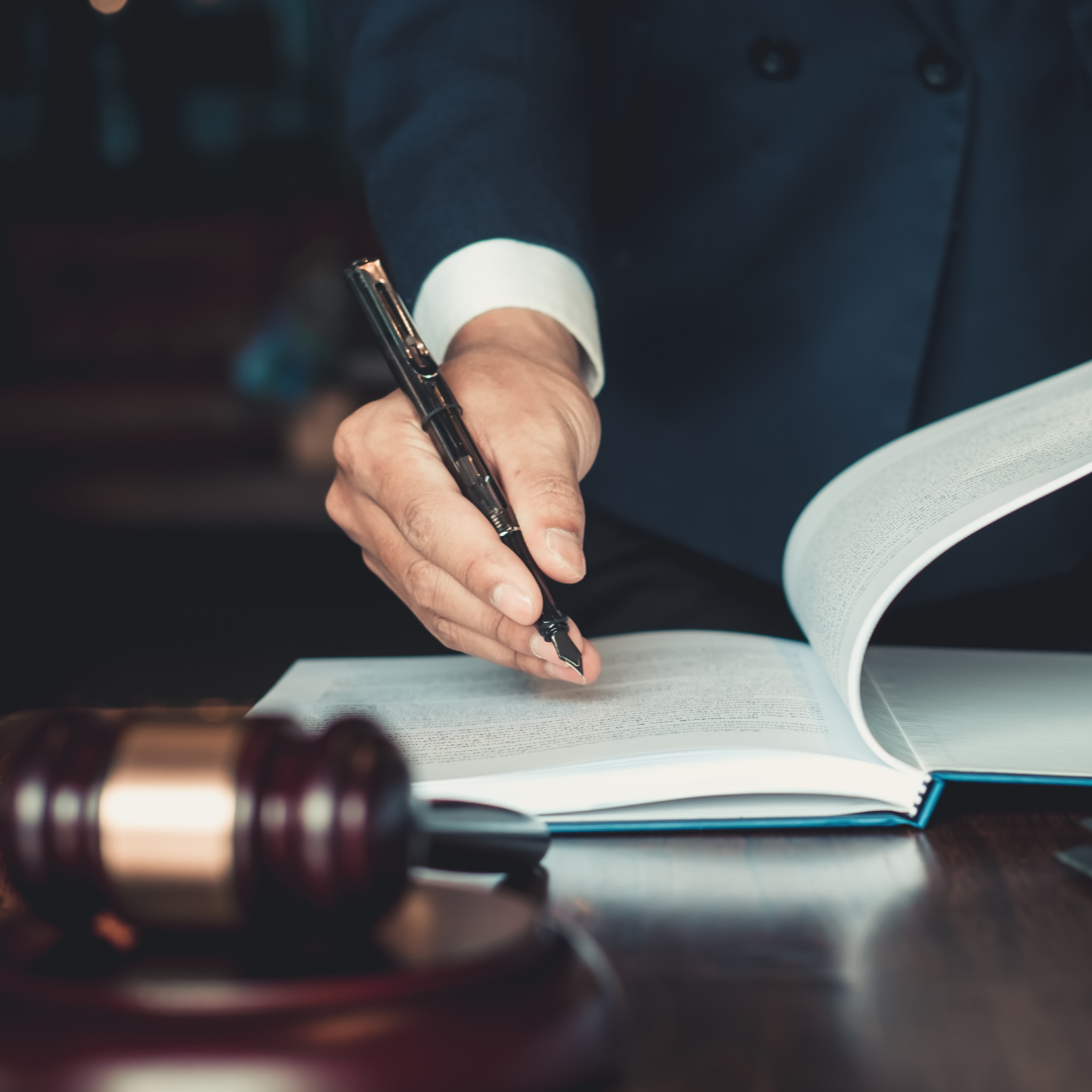 Person in suit signing a document, next to a gavel on a wooden table.