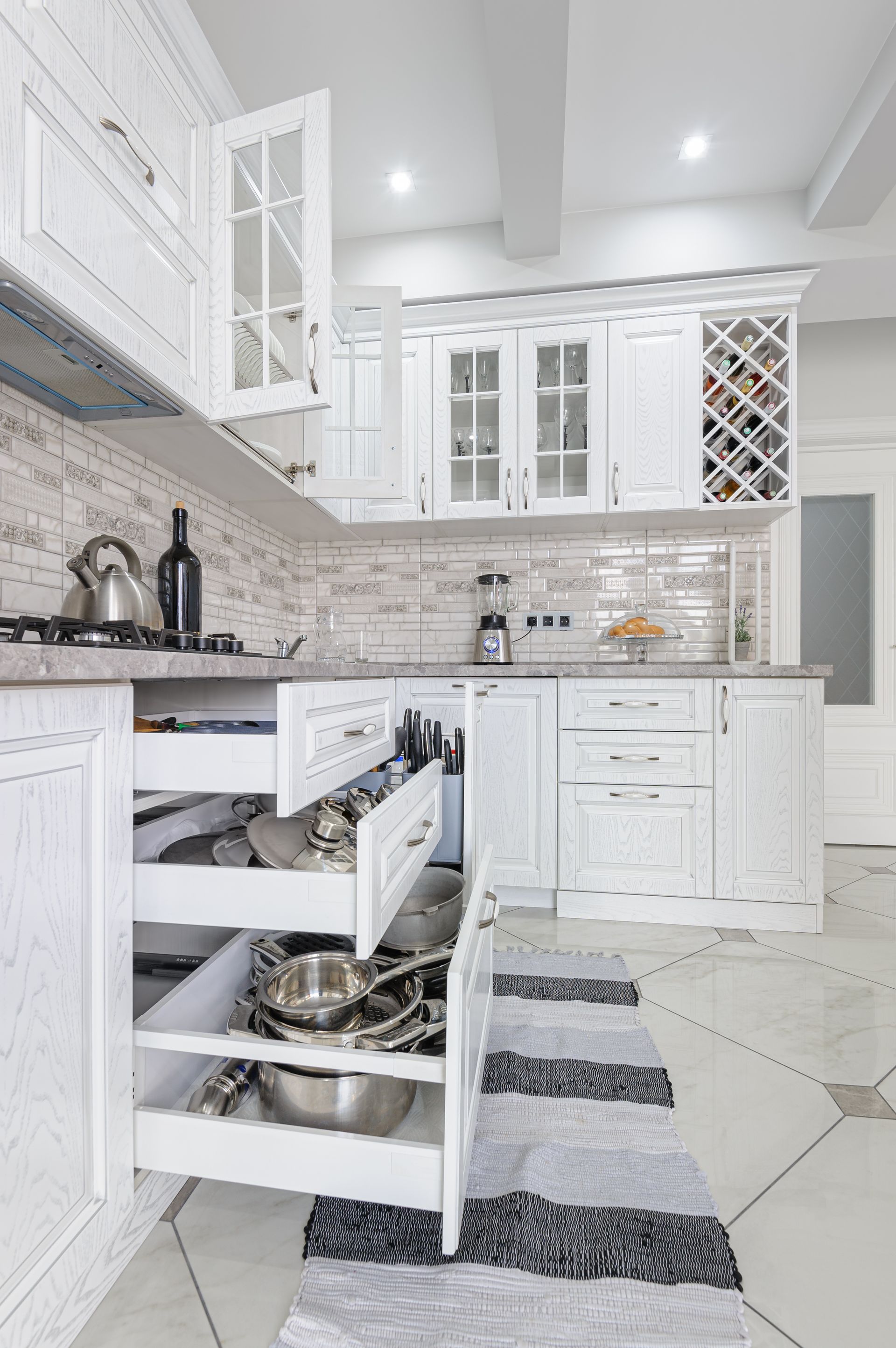 White kitchen with open drawers displaying utensils, pots, and pans. A wine rack and glass-fronted cabinets sit above a countertop.