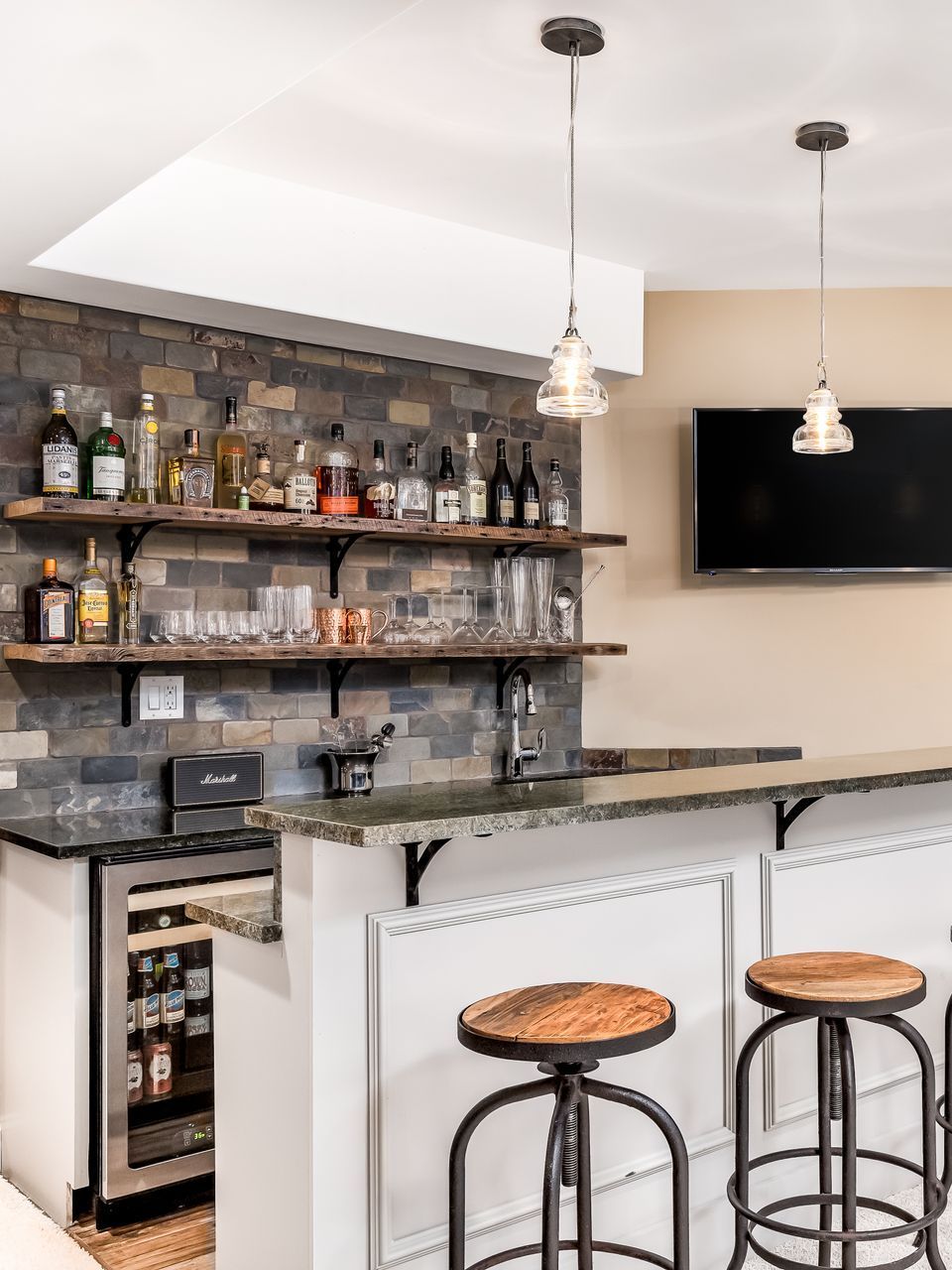 Cozy home bar with stone accent wall, floating shelves of liquor, countertop, and two bar stools.  A mini fridge is on the left.