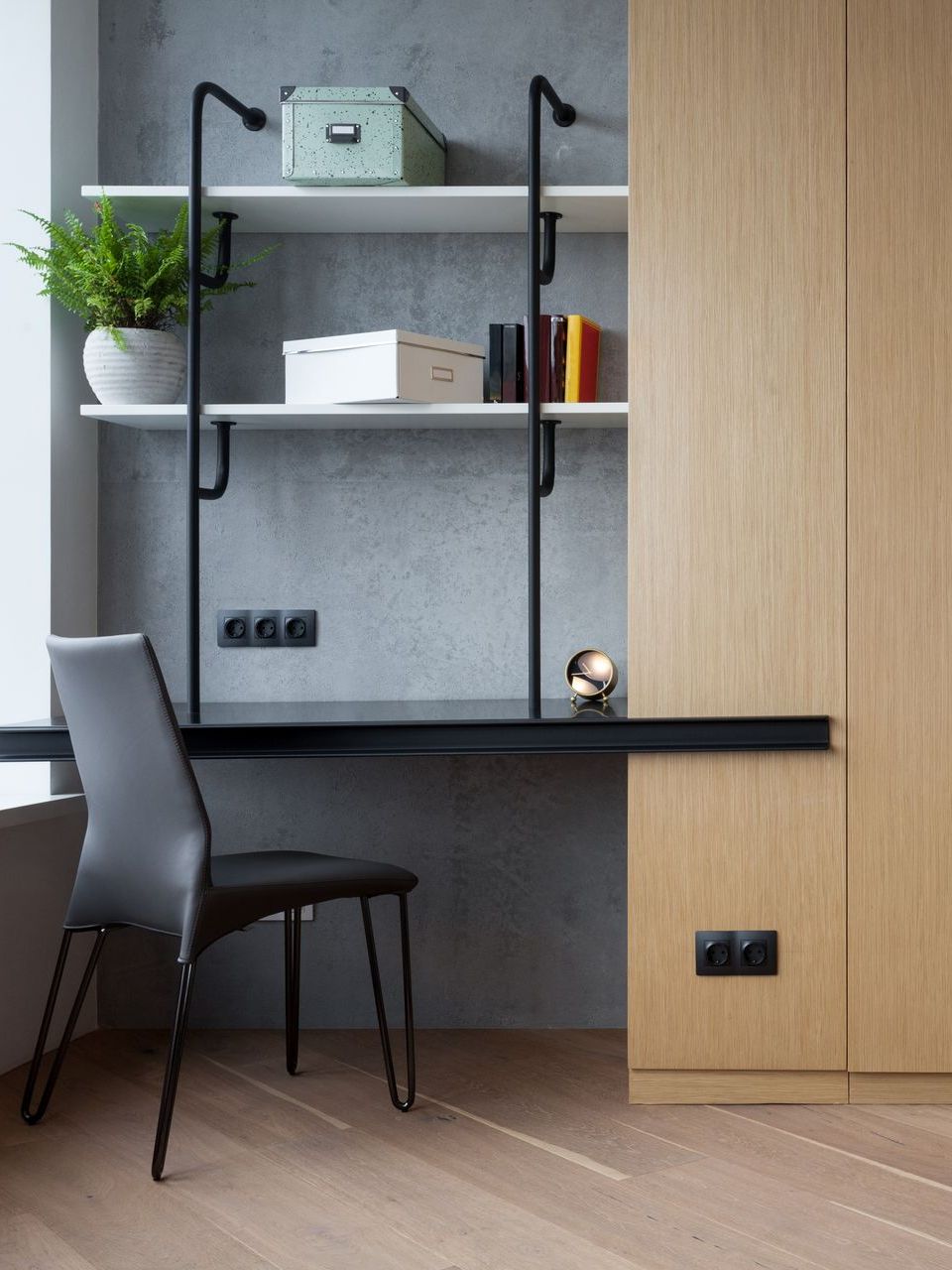 Modern office space with wooden cabinetry, black desk and chair, and a gray sofa. Light wood flooring, gray wall, and open shelving with plants.