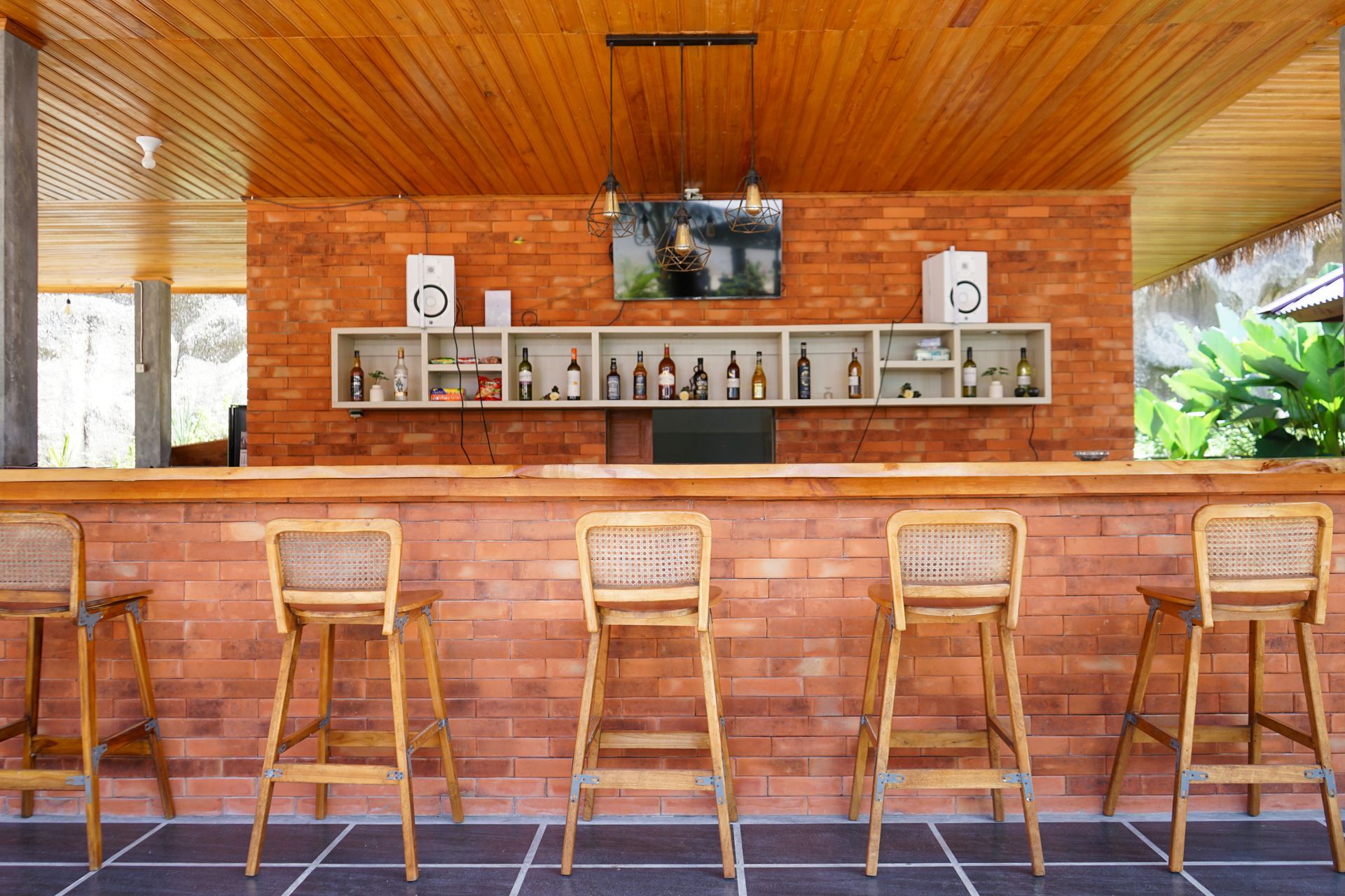 A bar with wooden stools and brick walls. A shelf displays bottles, speakers, and a TV, with a wooden ceiling overhead.