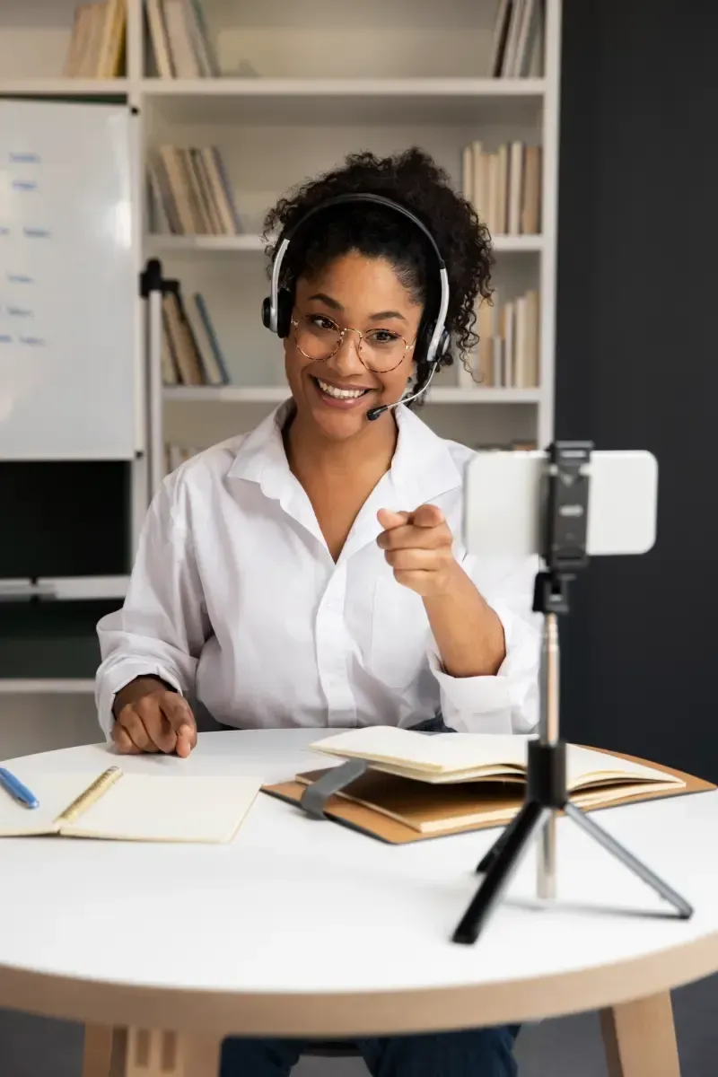 Smiling woman with headset, pointing at camera during video call, seated at desk with books.