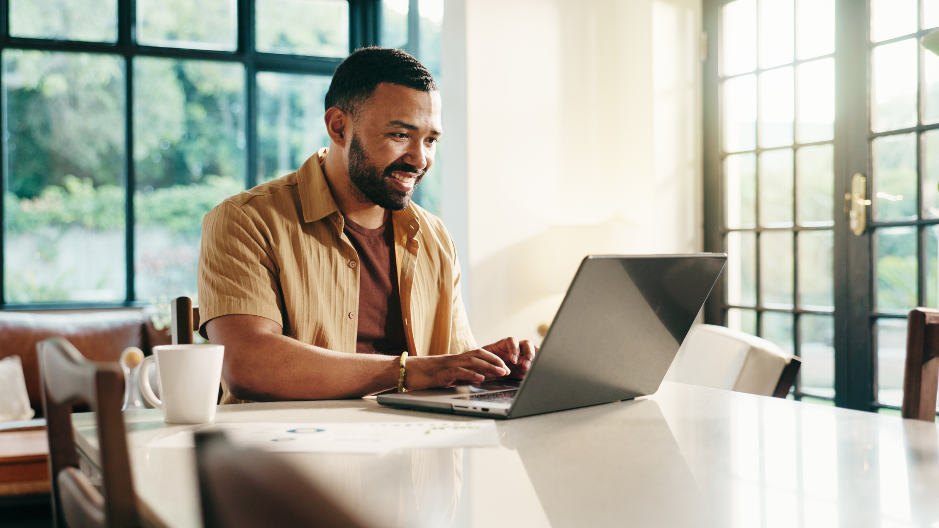 Man working on laptop at table, smiling. Coffee mug beside him, bright room.