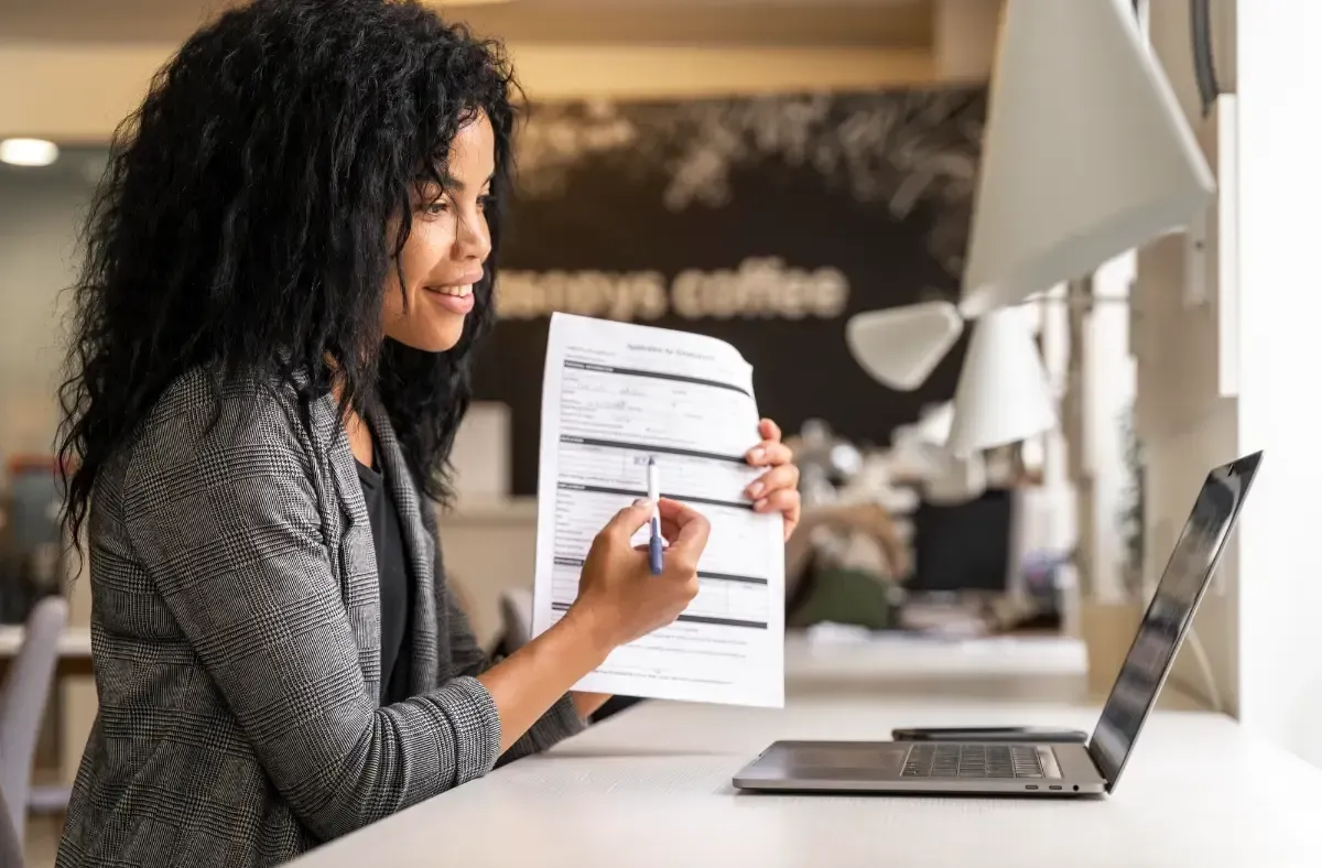 Woman with dark curly hair in a video call, pointing at document, laptop on desk near window.