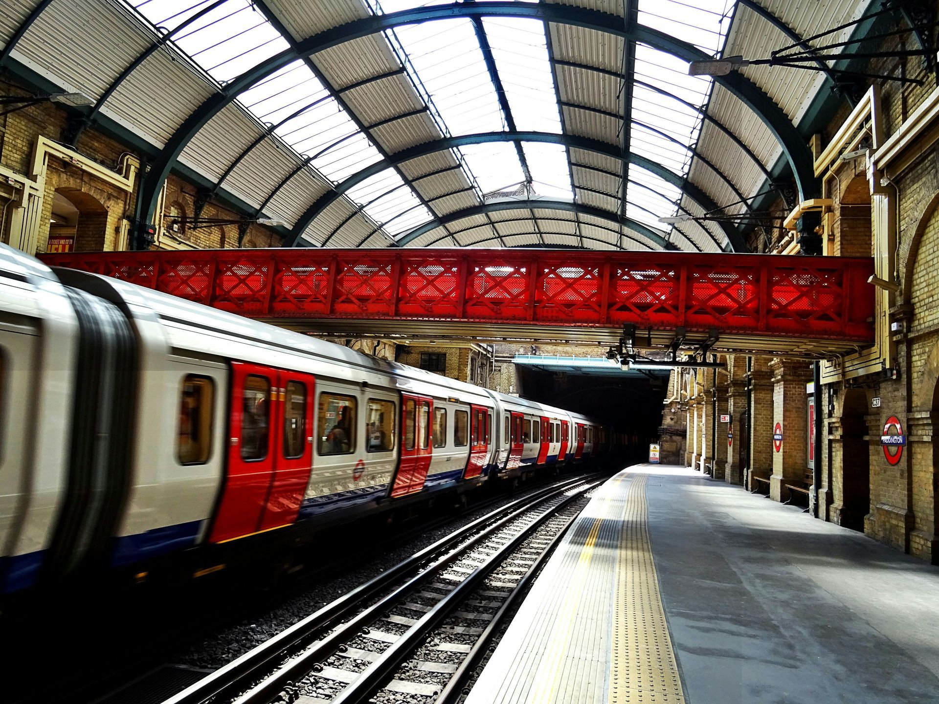 A red and white subway train entering a station tunnel. The station has a red bridge and arched ceiling.