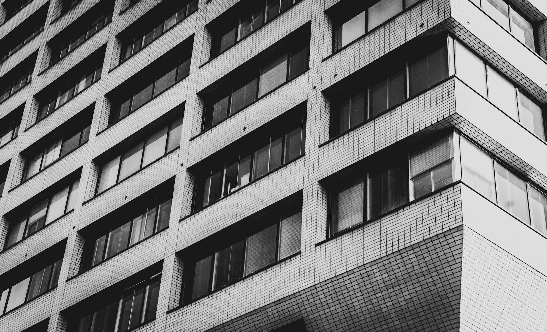Black and white view of a tall building exterior, angled upwards. Rows of windows and textured facade are visible.