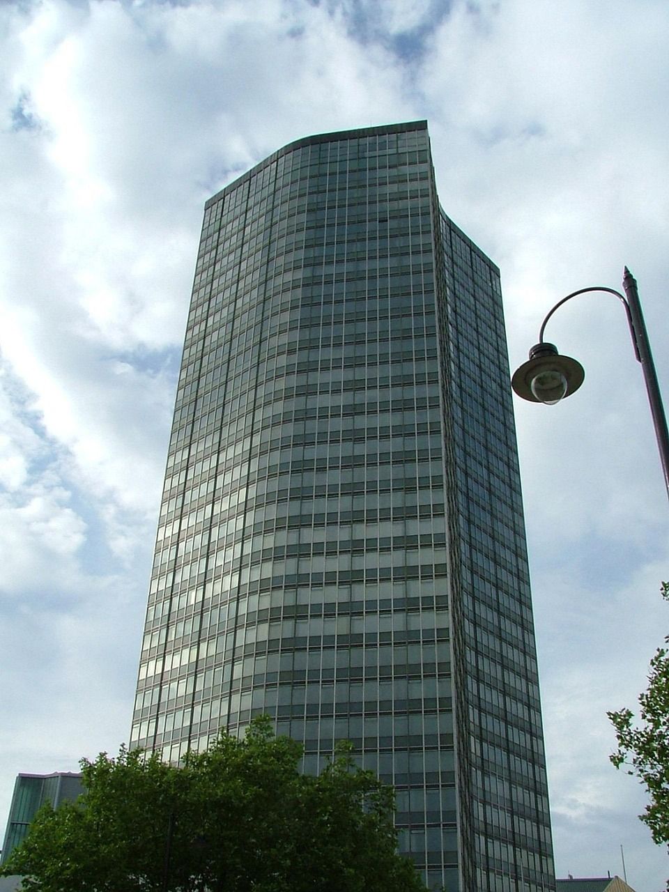 Modern, curved glass skyscraper against a cloudy sky, with a street lamp in the foreground.