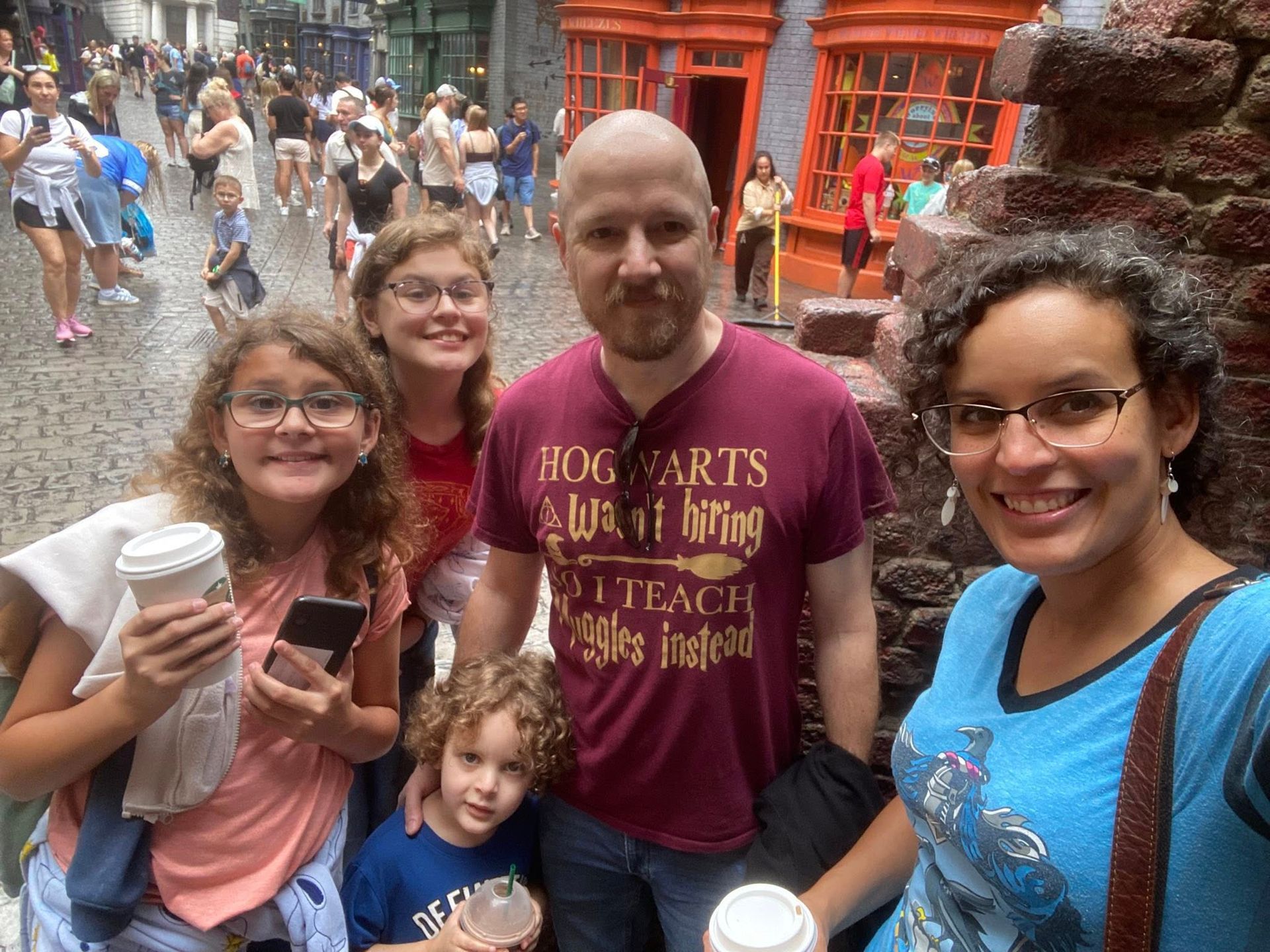 Family in a wizard-themed setting, smiling. People in the background. Buildings are orange. People holding drinks.