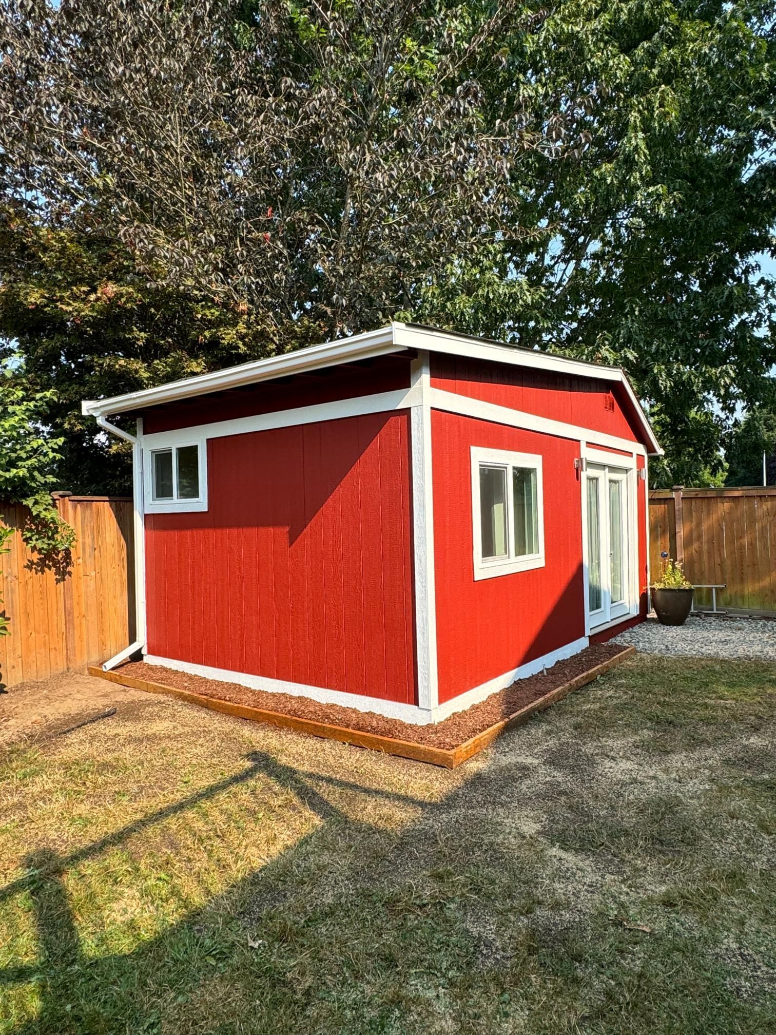 Red shed with white trim, two windows, and glass doors, set in a grassy yard, near a fence and trees.