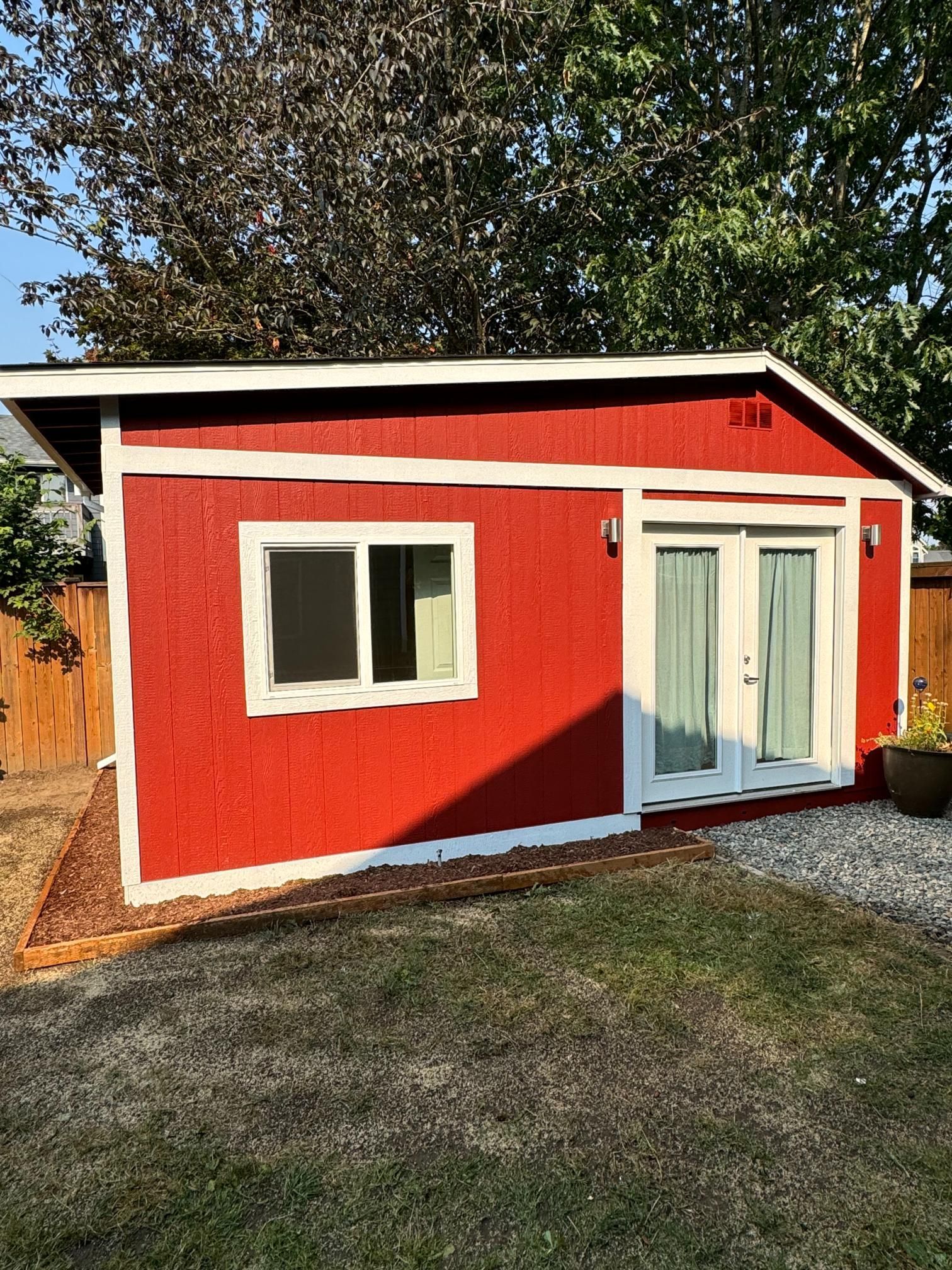 Red shed with white trim, window, and French doors in a grassy backyard, surrounded by trees and a fence.