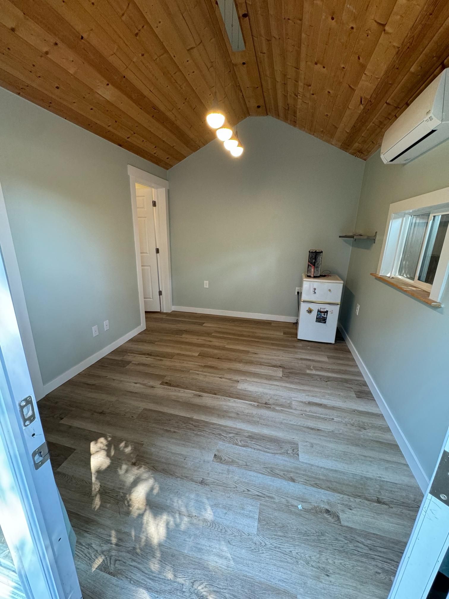 Room with wood ceiling, light blue walls, and wood-look flooring. A/C unit, white cabinet, and a door are visible.