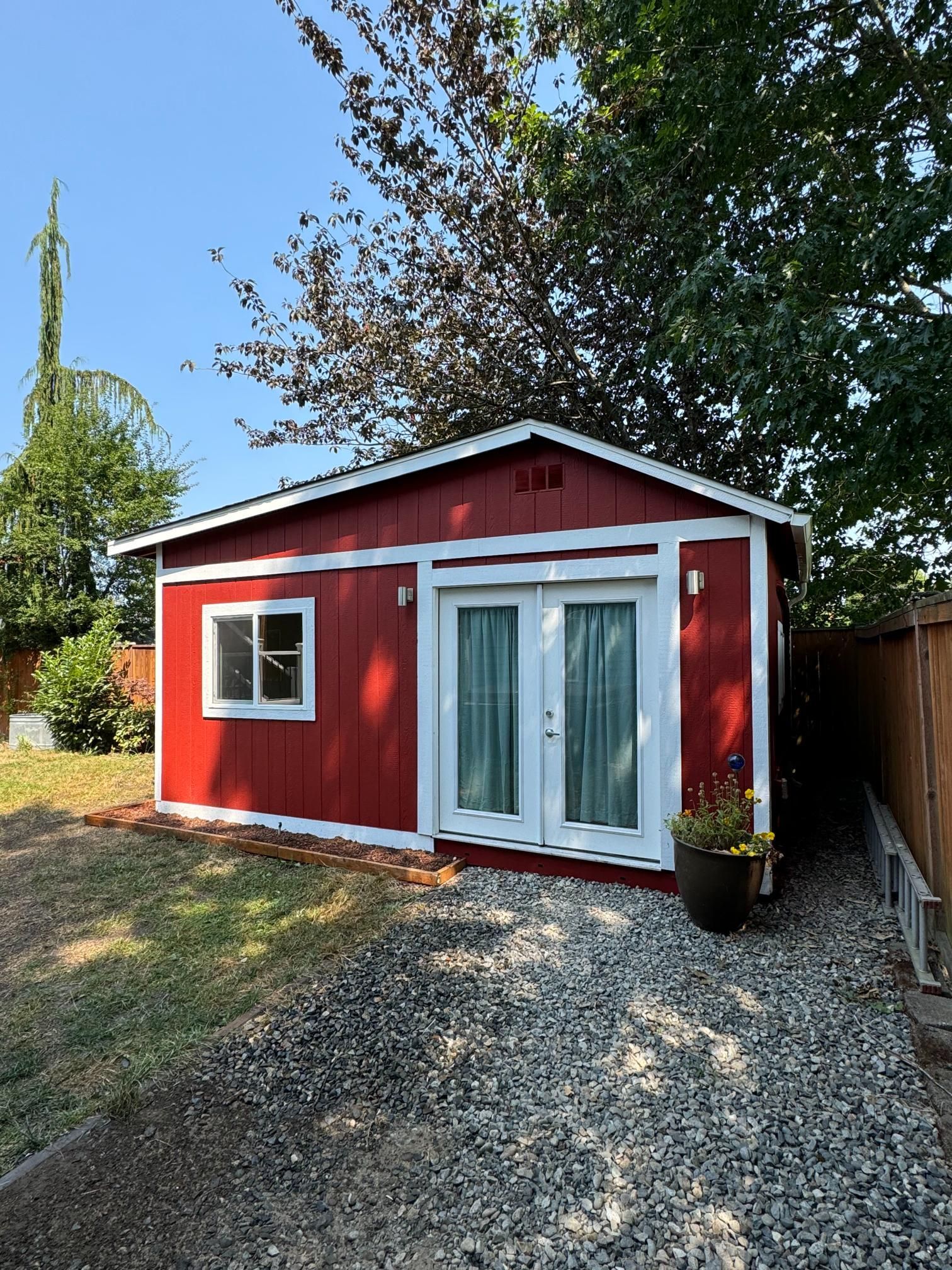 Red shed with white trim, double doors, and window, gravel path, surrounded by trees and lawn.