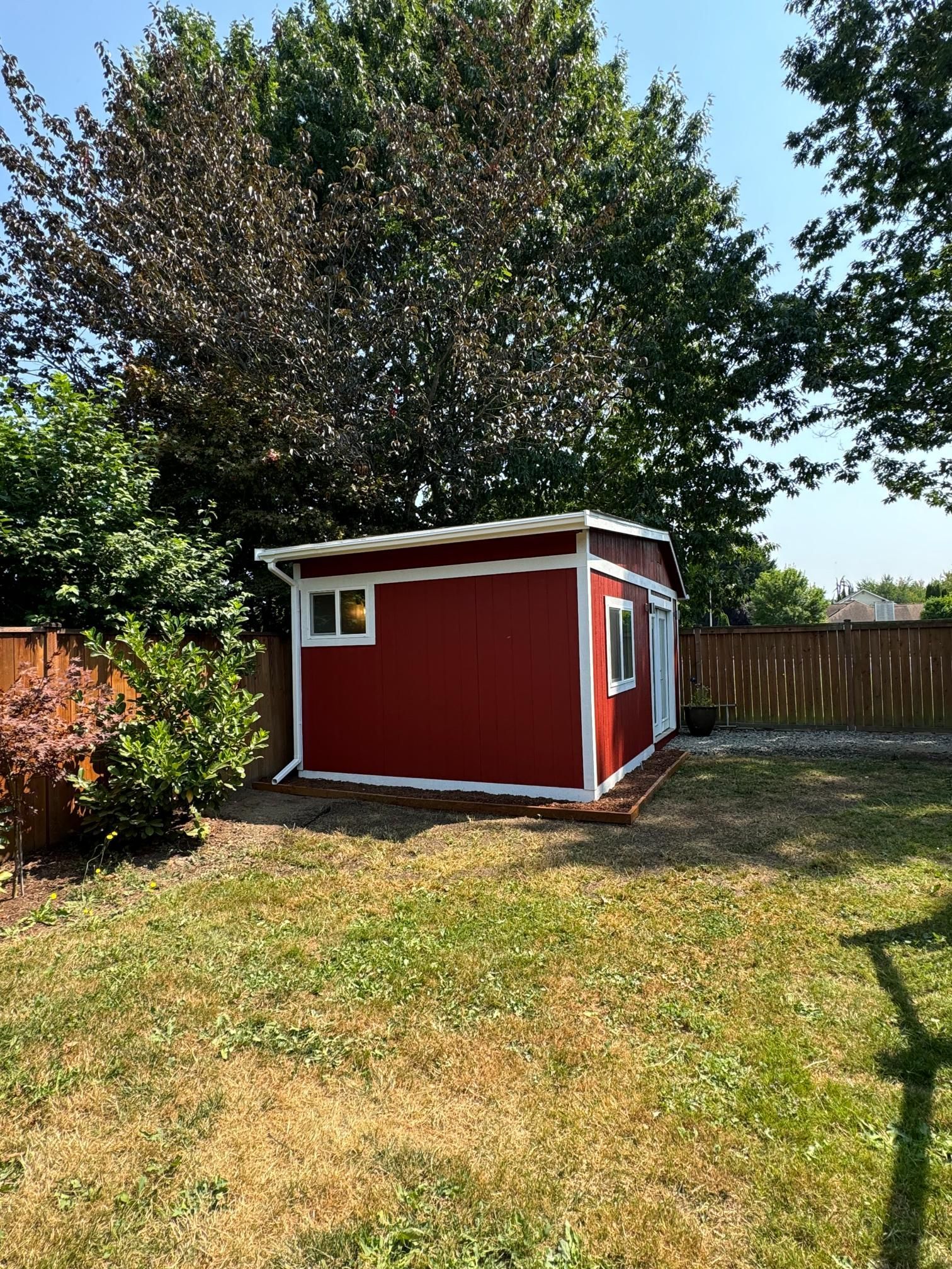 Red shed with white trim in a grassy backyard. Two windows, a door, and trees are present.