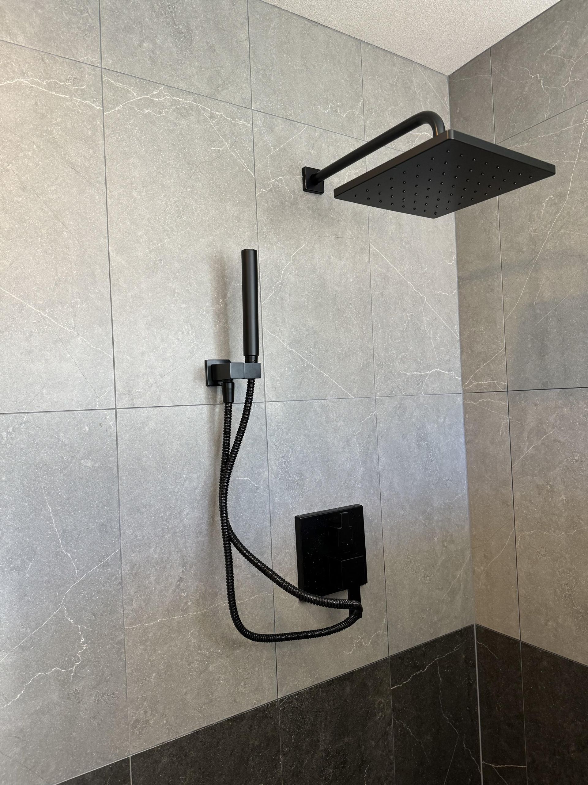 Black shower fixtures on gray tiled wall. Square overhead shower and handheld shower head.