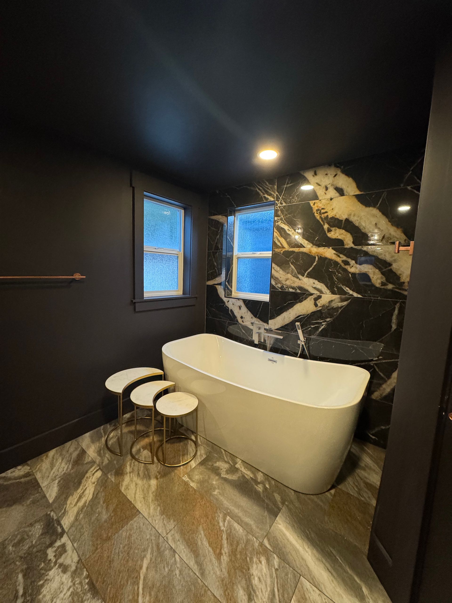 Bathroom with freestanding tub, black and gold tiled accent wall, and marble-look flooring.