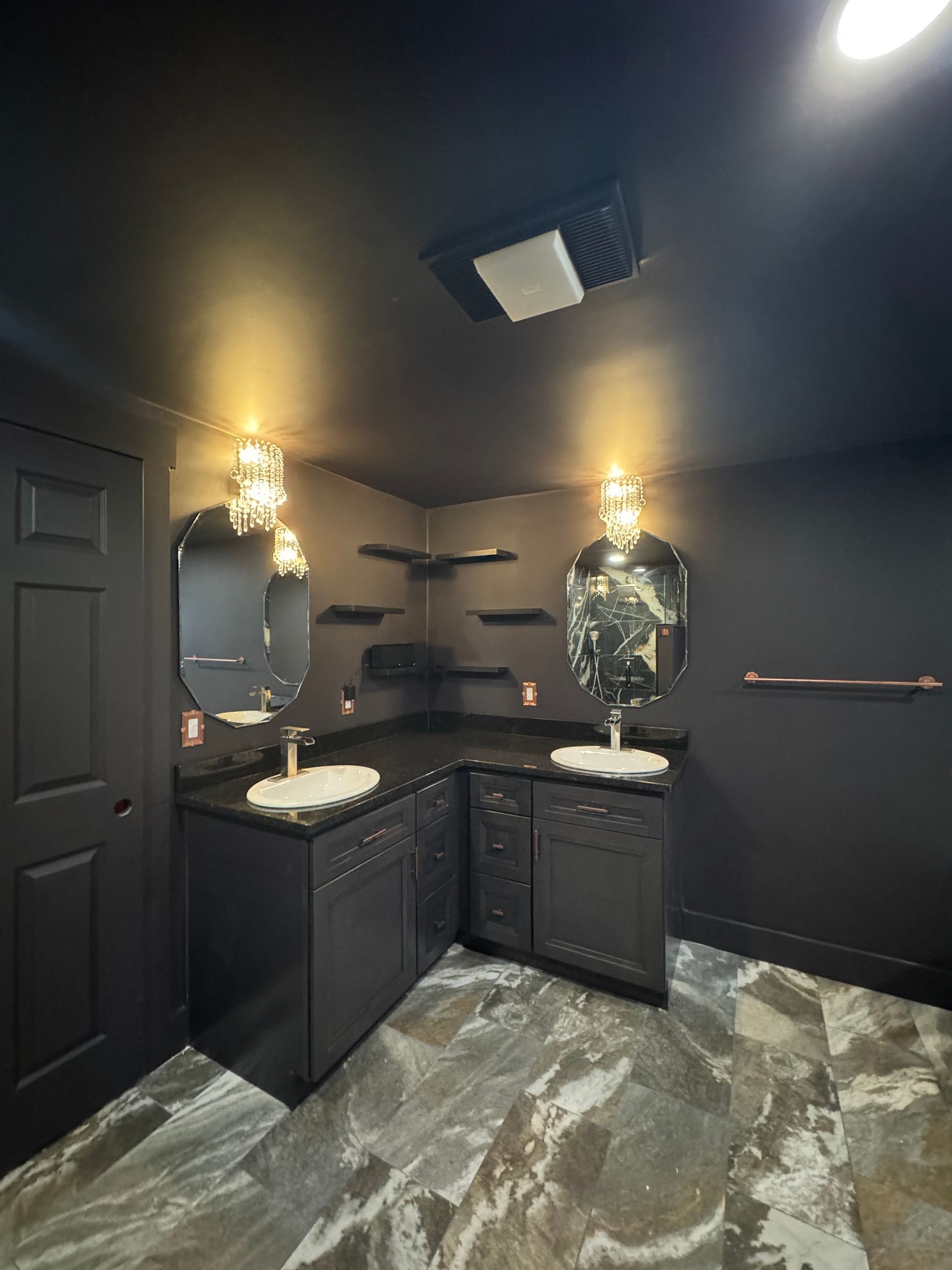 Black bathroom with two sinks, dark cabinets, a corner vanity, and a textured floor.