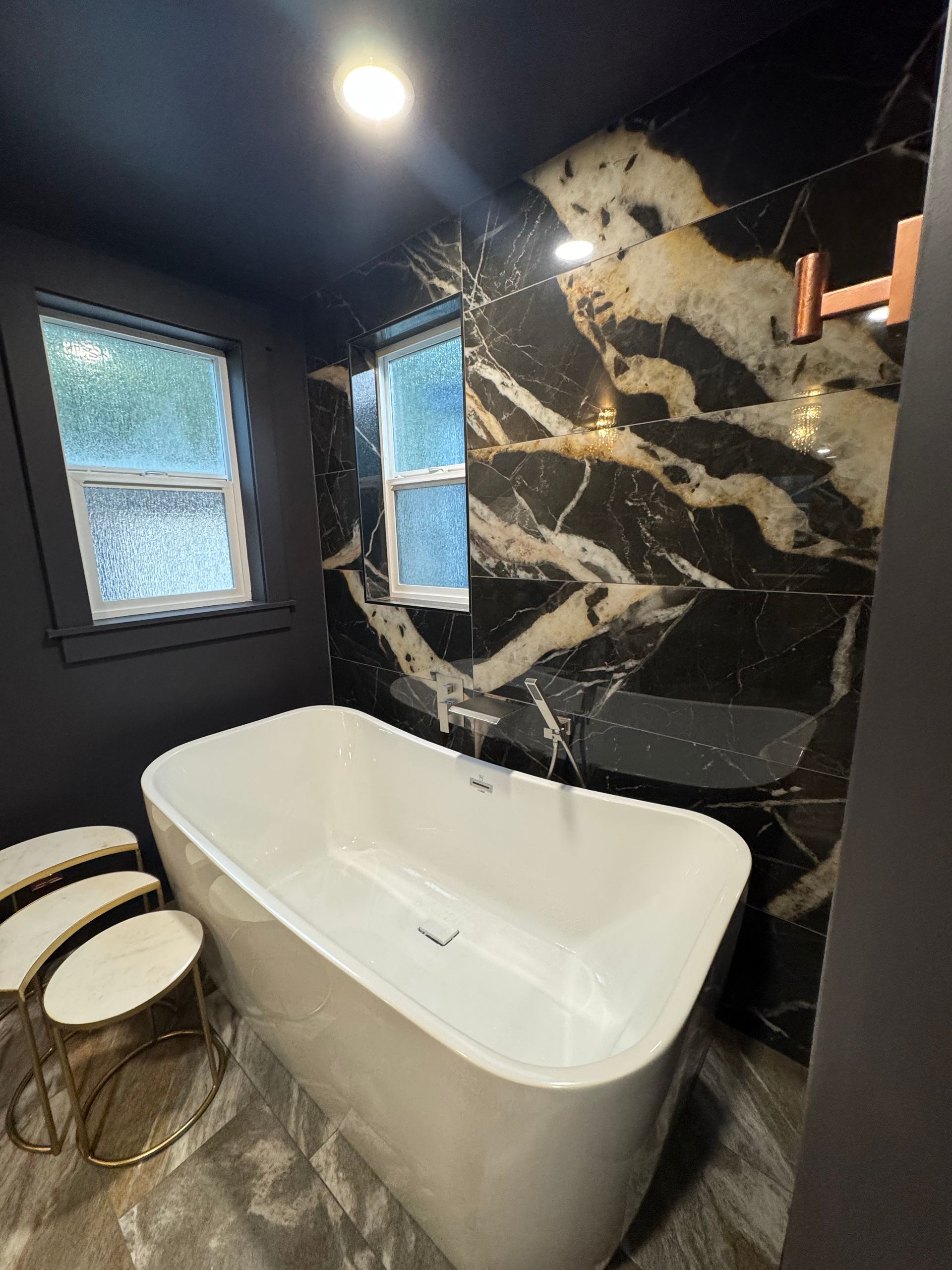 Modern bathroom with a white tub, black marble wall, and two windows.