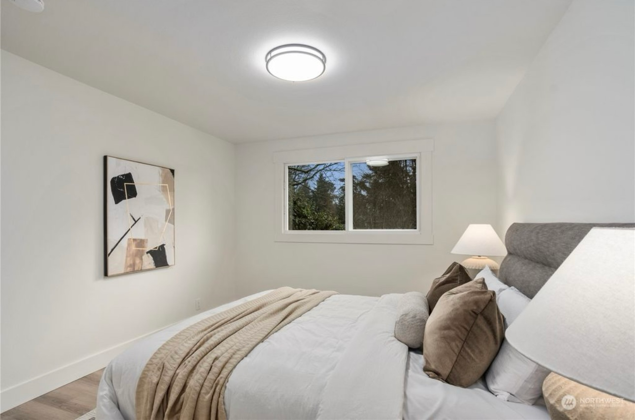 Bedroom with a white bed, beige throw, and pillows; window to trees; abstract art.