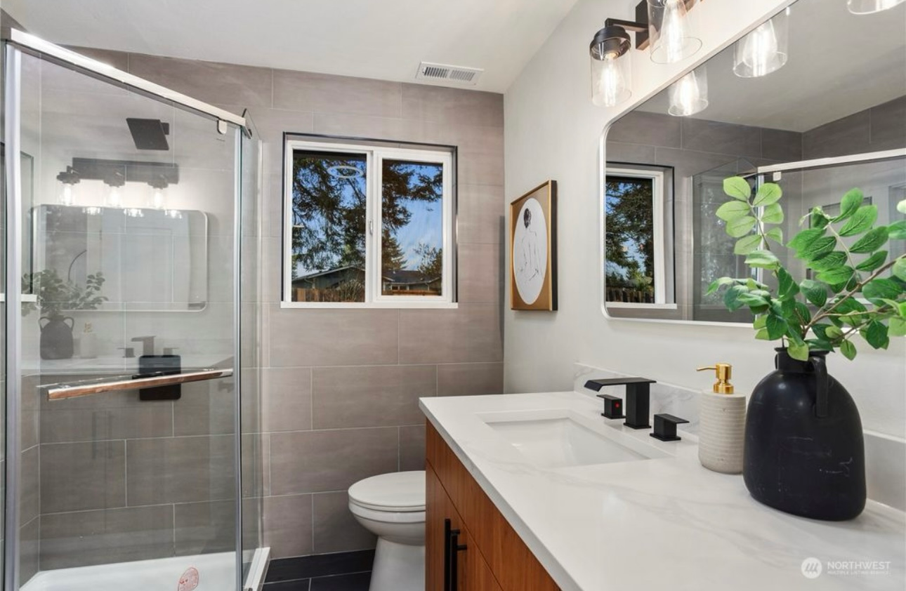 Modern bathroom with gray tile walls, glass shower, wood vanity, and decorative plants.