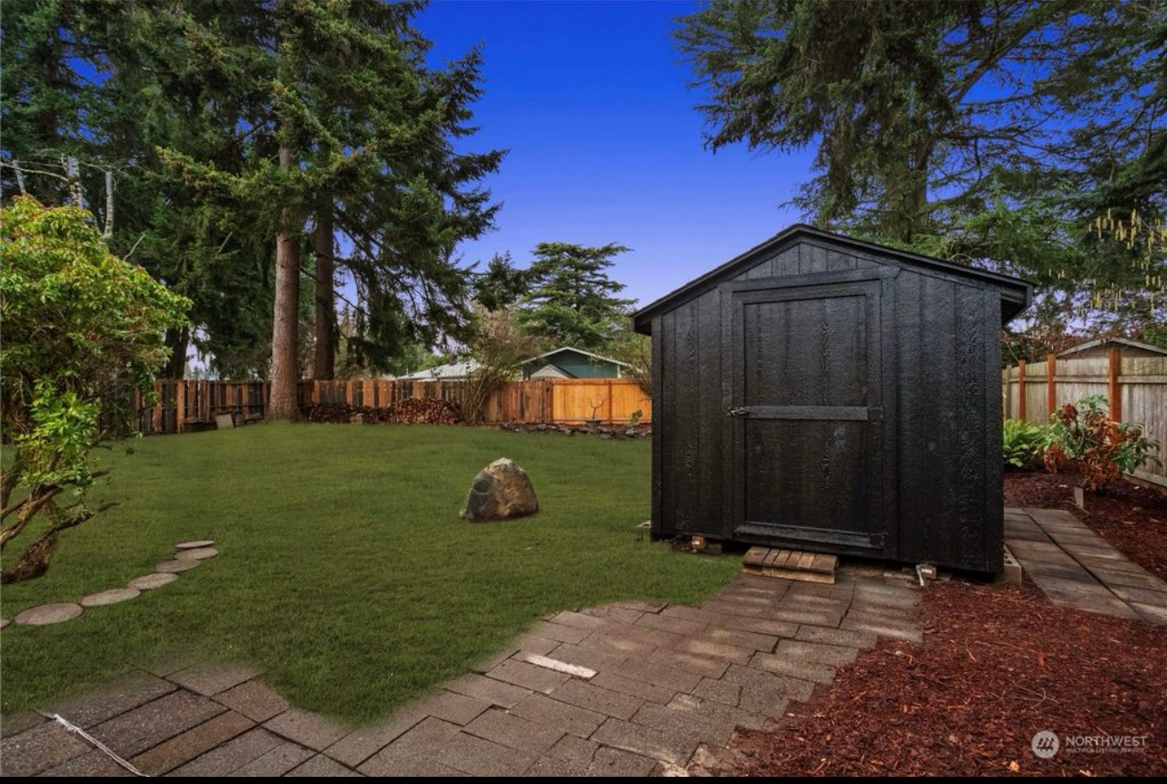 Black shed in backyard with green grass, trees, and blue sky. Stone pathway leads to shed door.