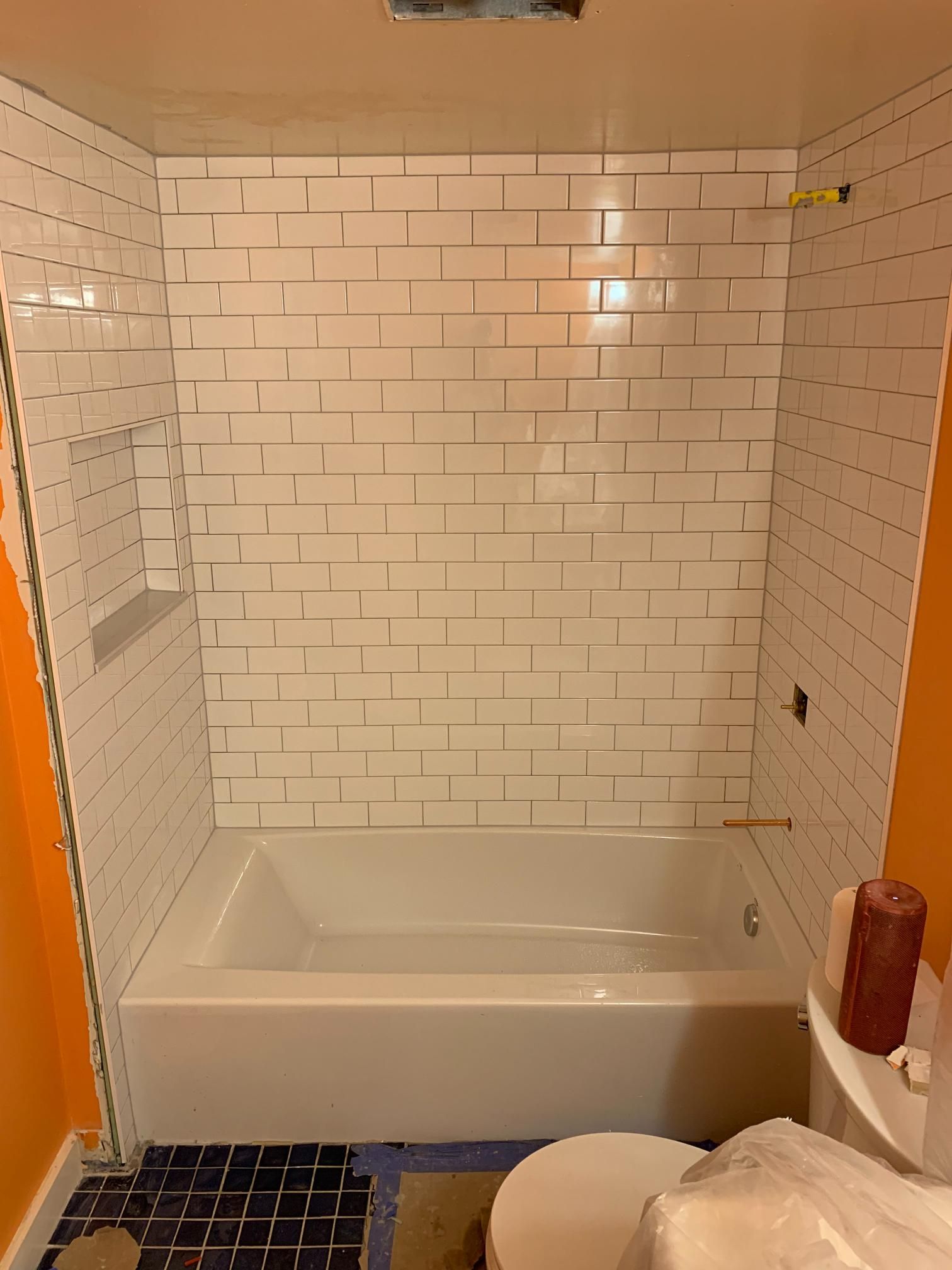 White tiled shower enclosure with built-in shelf, above a white bathtub, with orange wall and a toilet in the foreground.