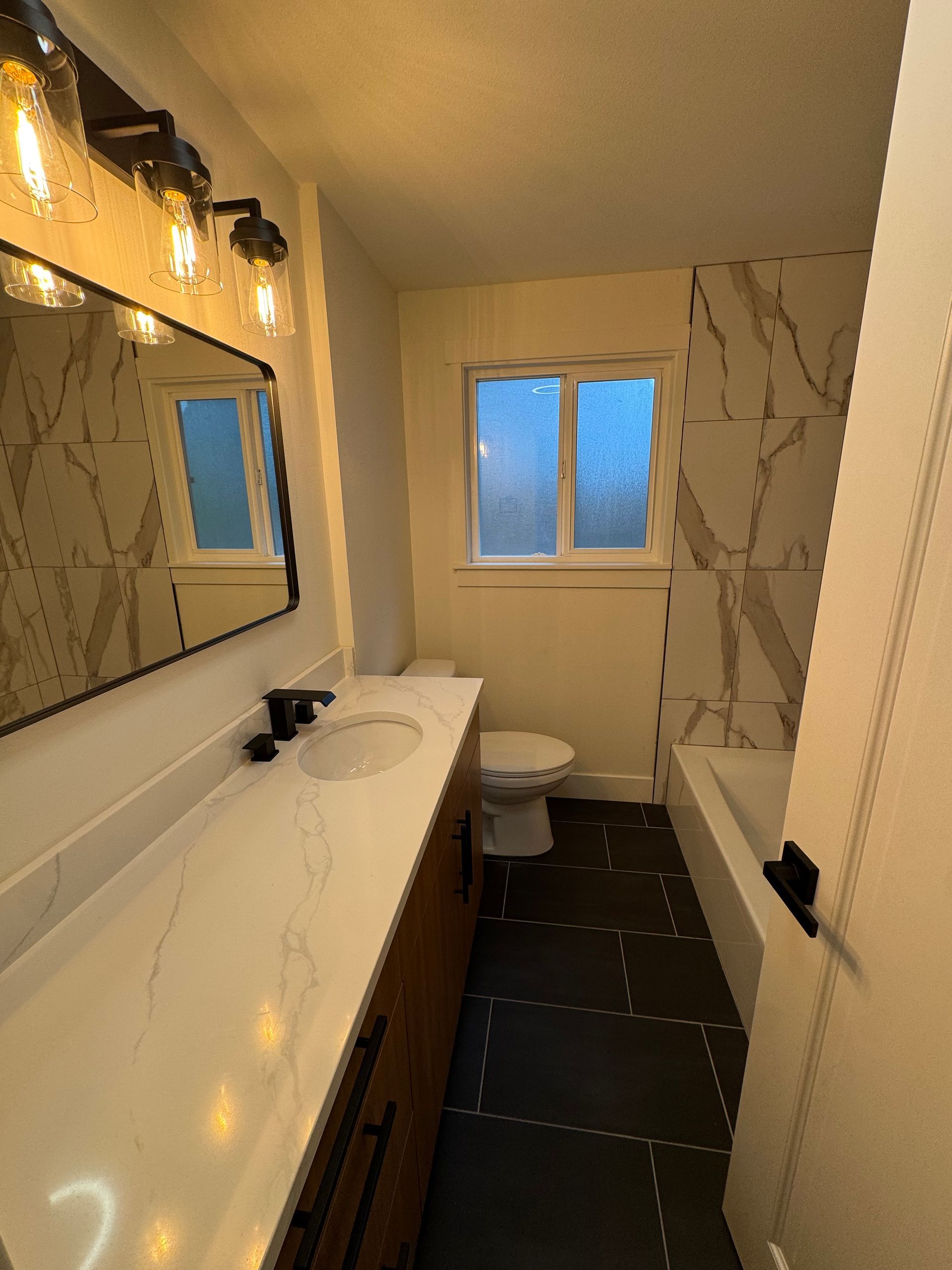 Bathroom with white marble-look tile, dark wood vanity, black fixtures, and a long mirror with exposed bulb lights.