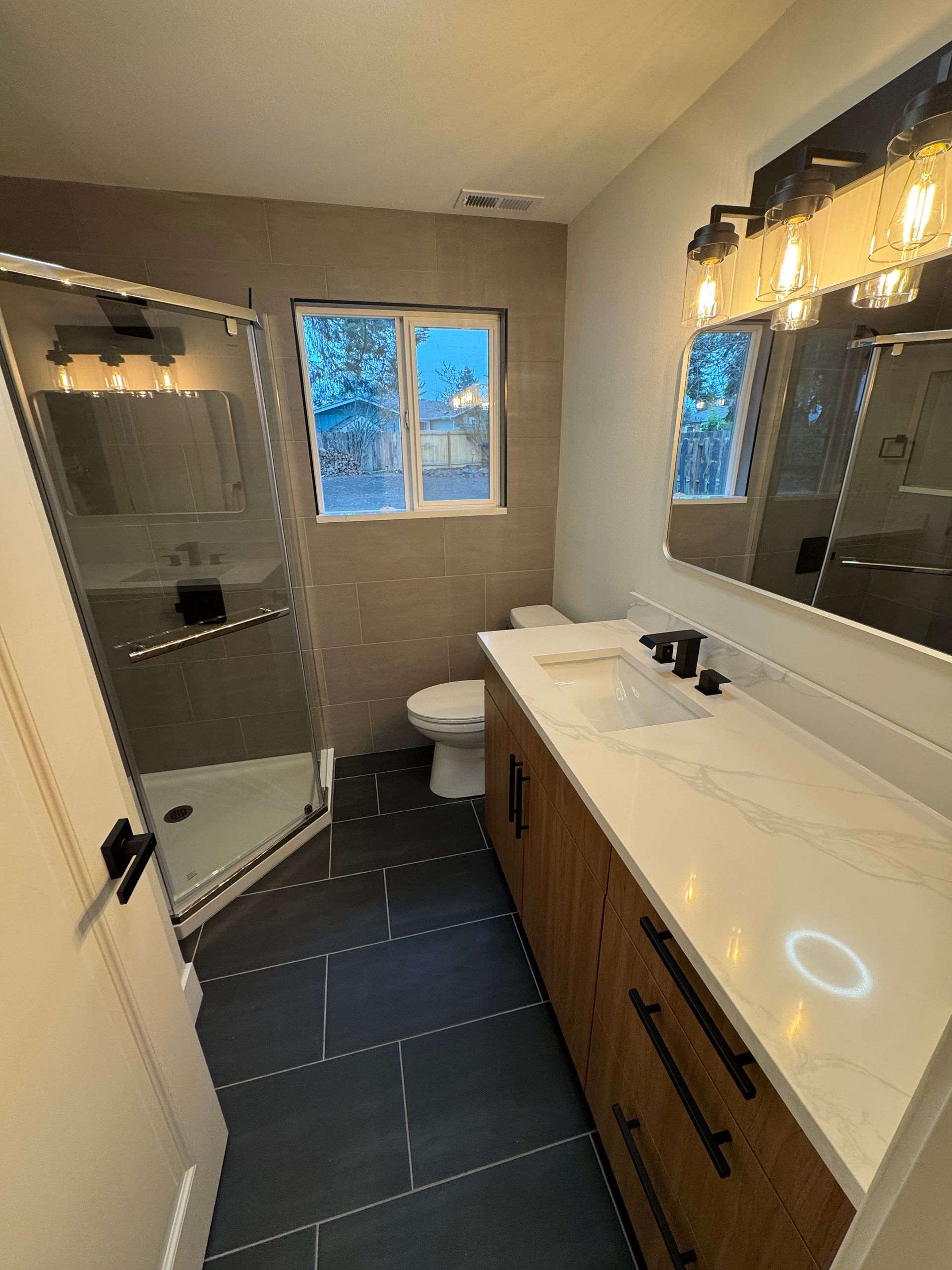 Modern bathroom with a wood vanity, white countertop, black fixtures, and a glass shower.