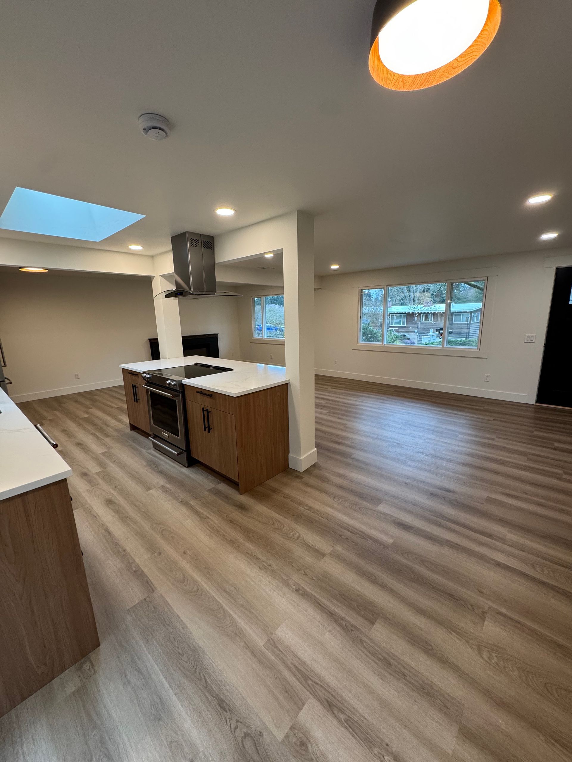 Open-plan kitchen and living area with wood-look flooring, island with stove, and natural light from windows and a skylight.
