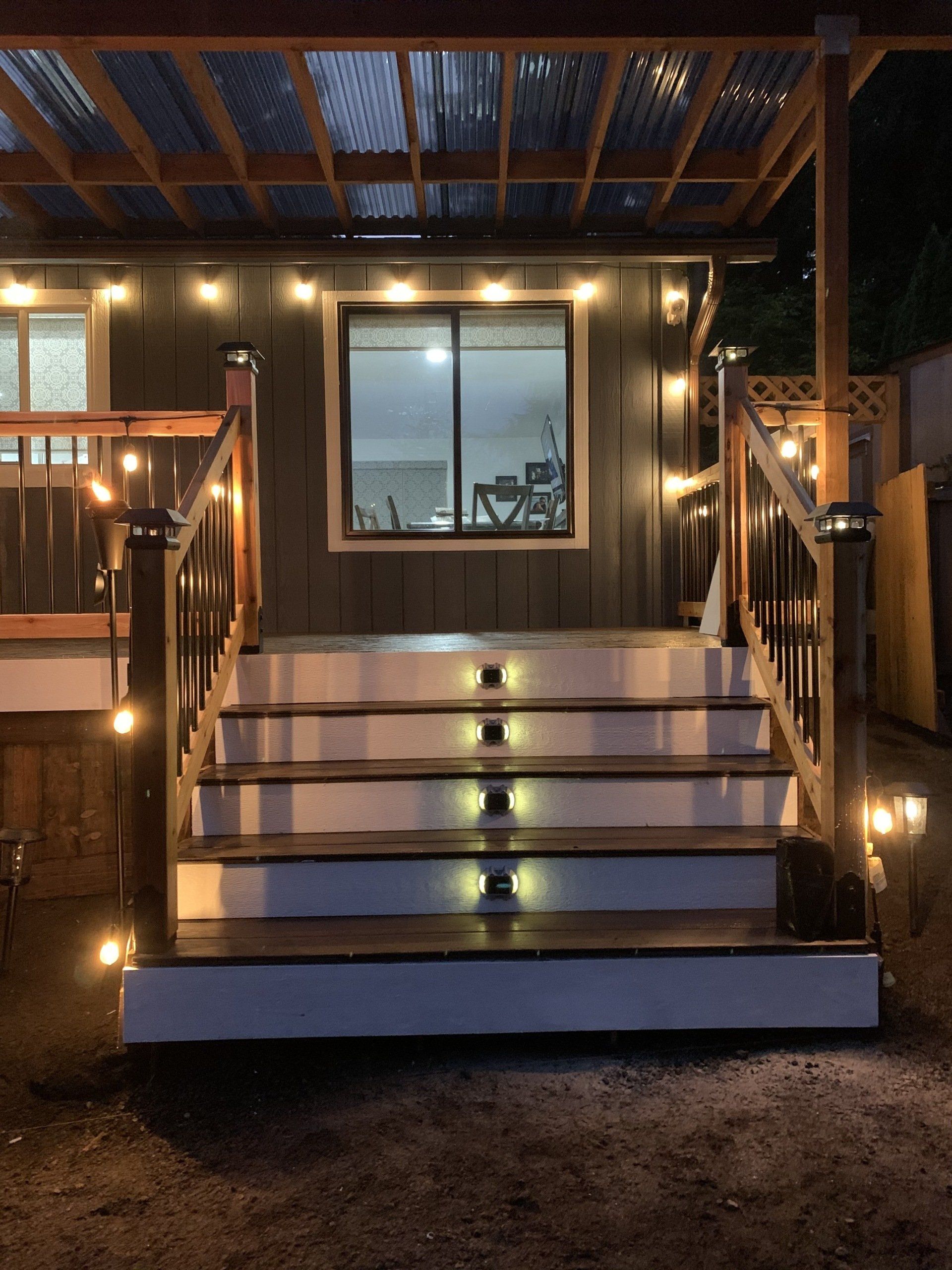 Wooden stairs lit with string lights lead to a building with a window; corrugated roof overhead.