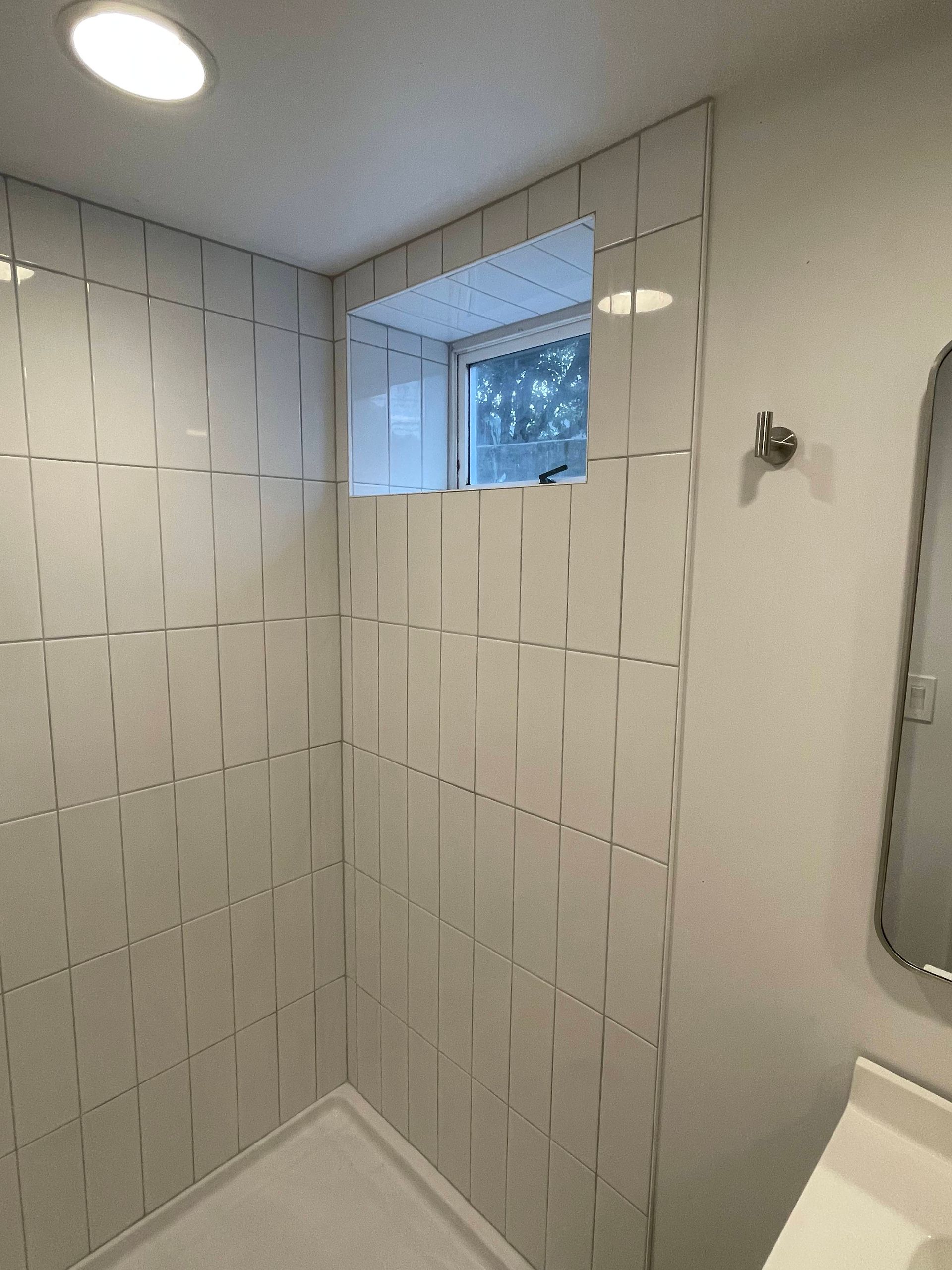 White-tiled shower with small window, recessed ceiling light. Towel hook on the wall.