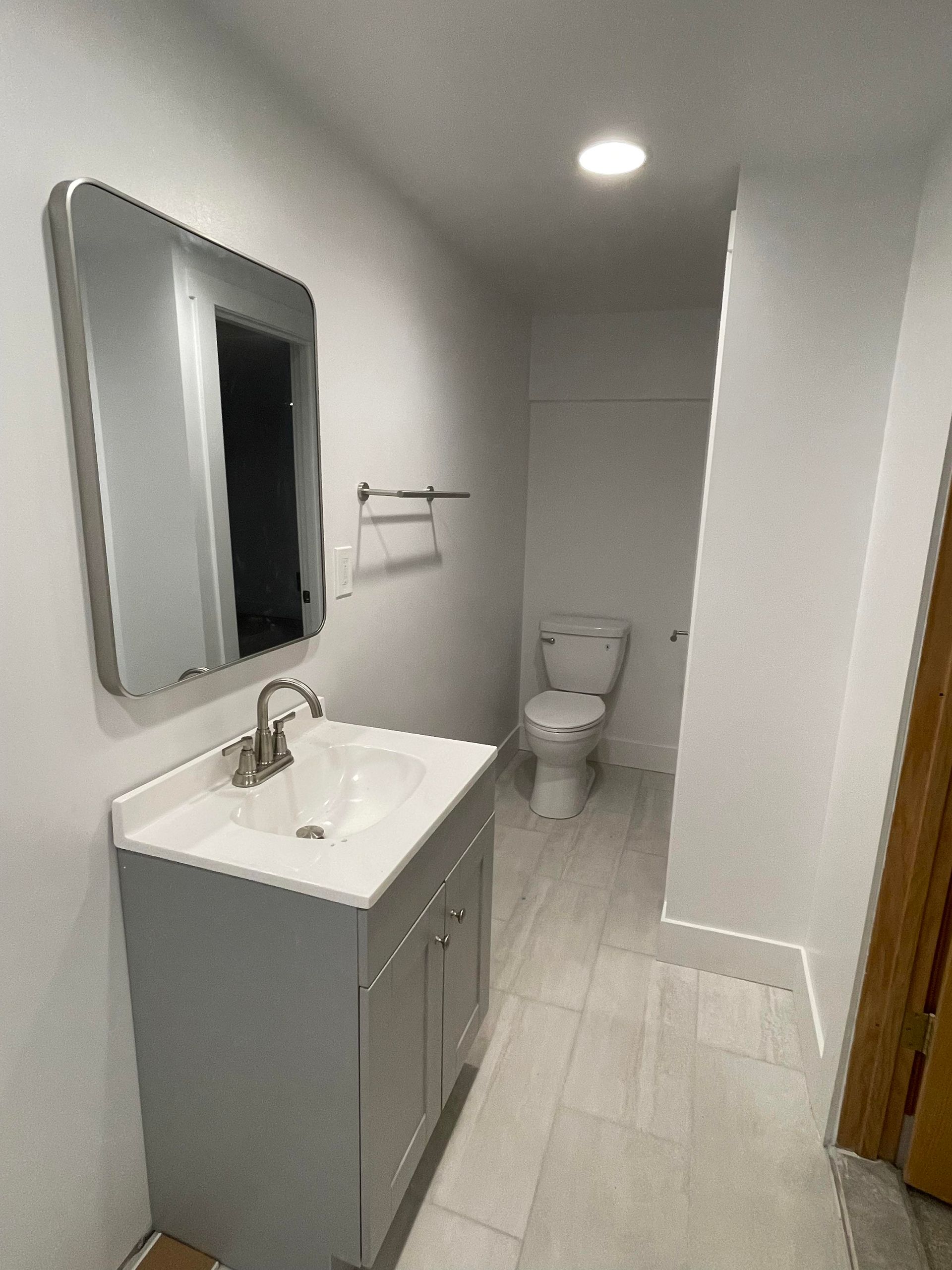 Bathroom with vanity, toilet, mirror, and towel rack. White walls, gray vanity, and light floor tiles.