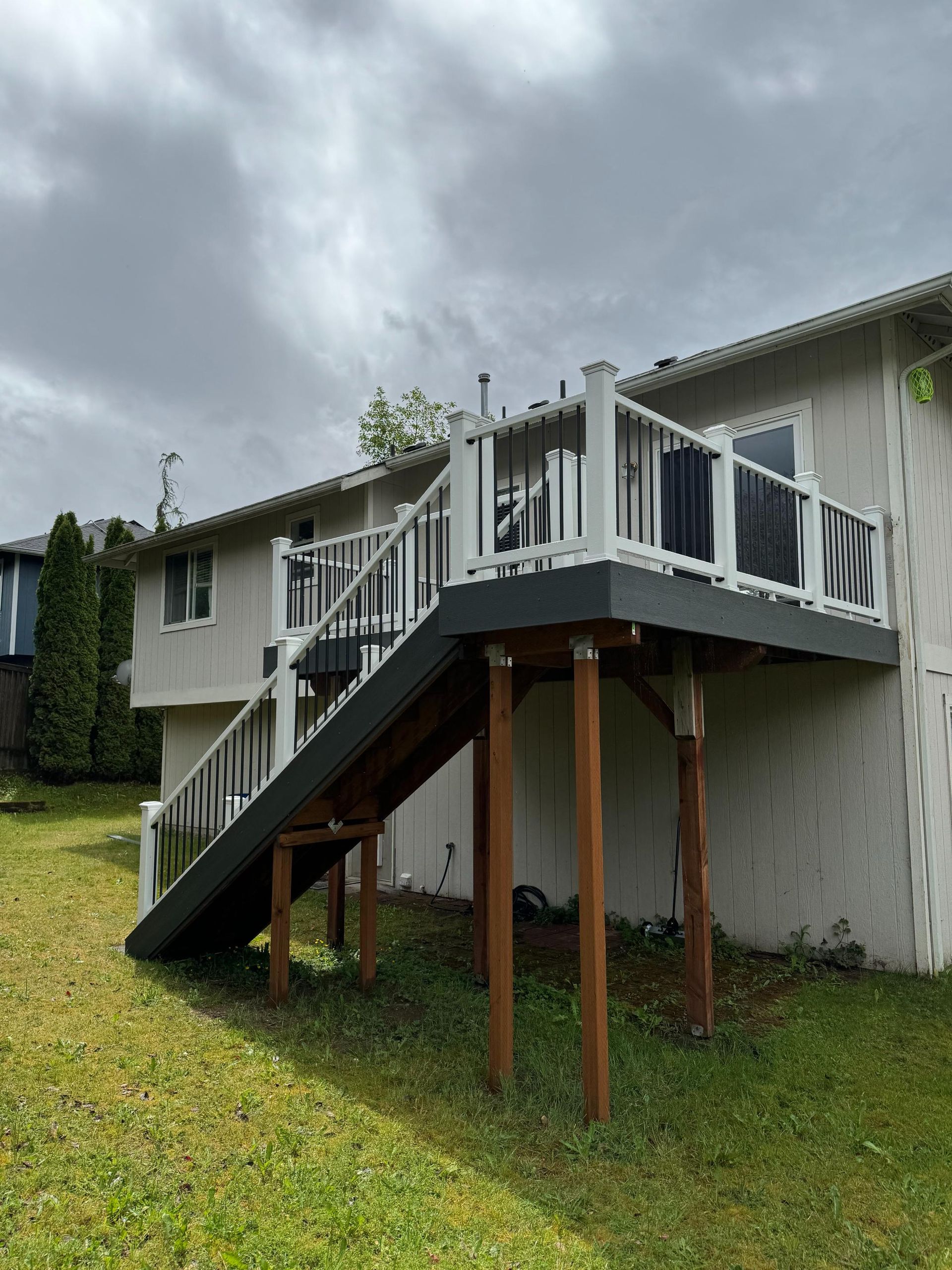 Exterior view of a deck with white railings and gray decking; supported by wooden posts, against a light-colored house.