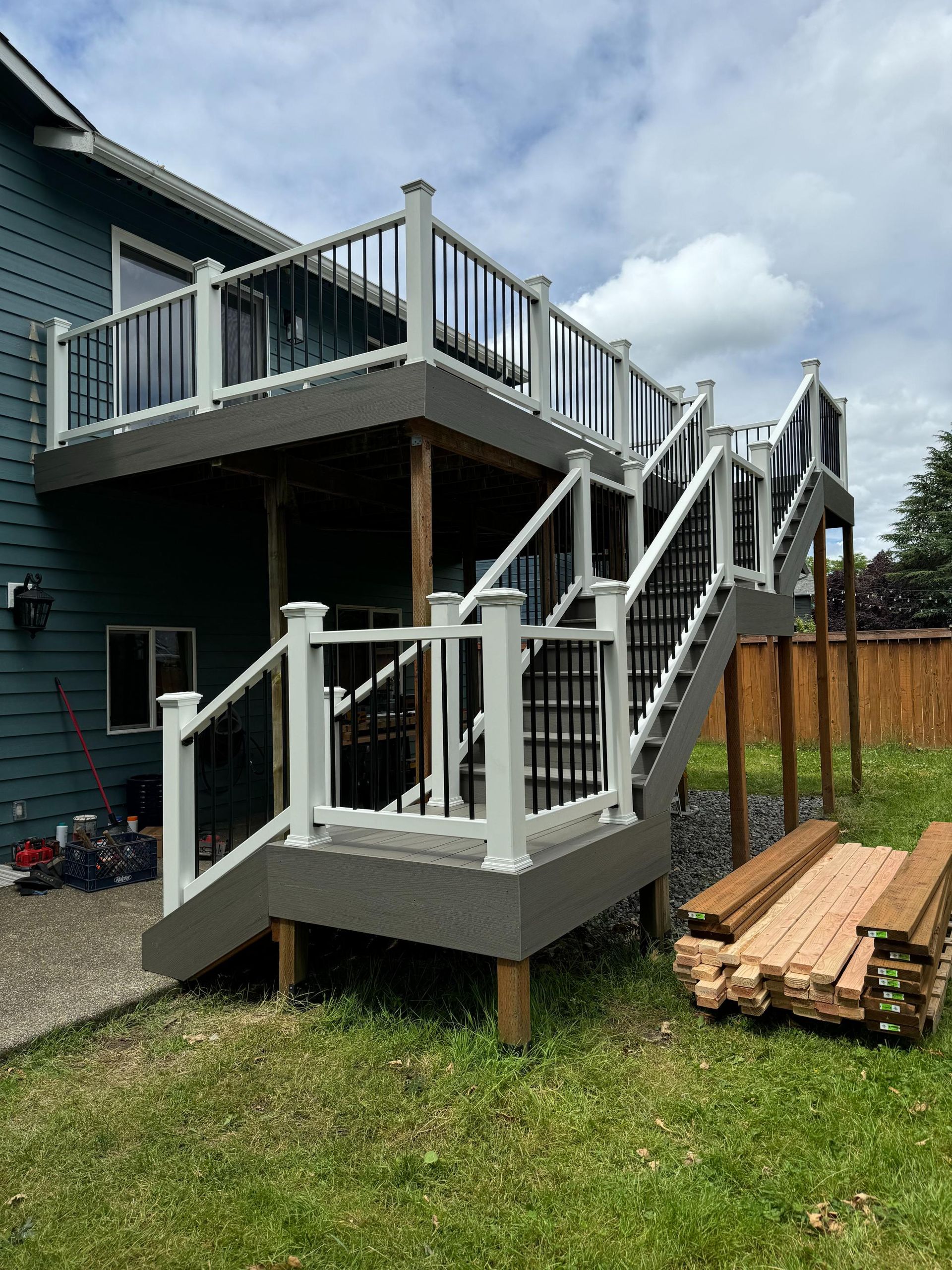 Multi-level deck with white and black railings, gray decking, and wooden supports, next to a green house.