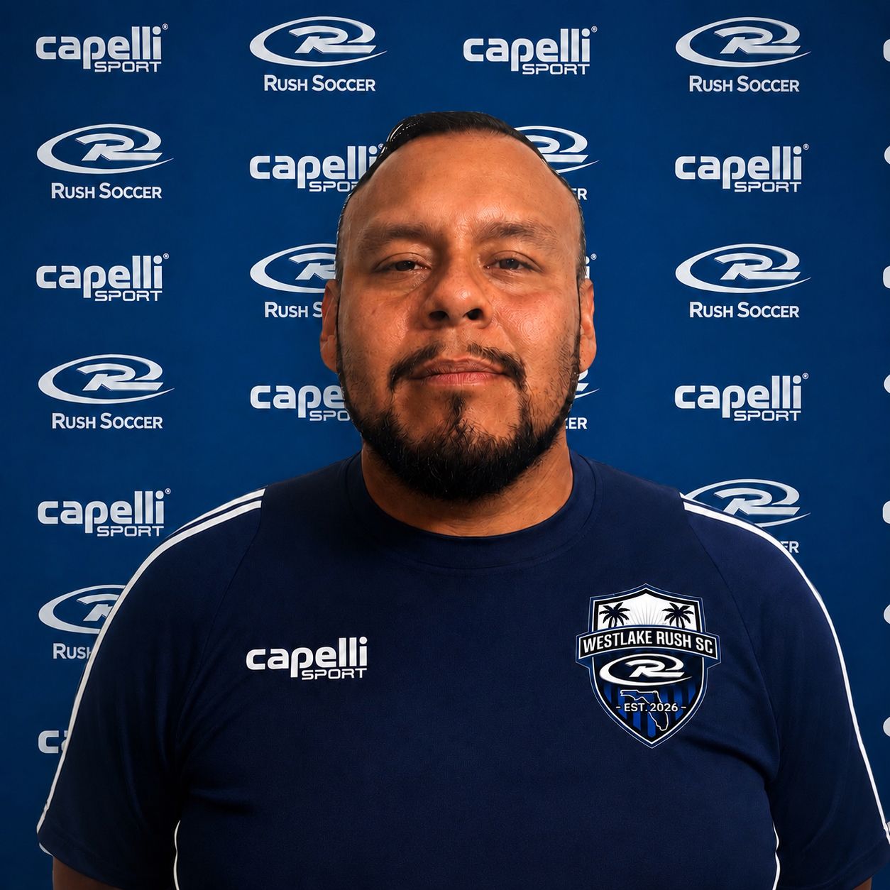 Man standing in front of a blue sponsor backdrop, wearing a navy sports shirt and smiling slightly.