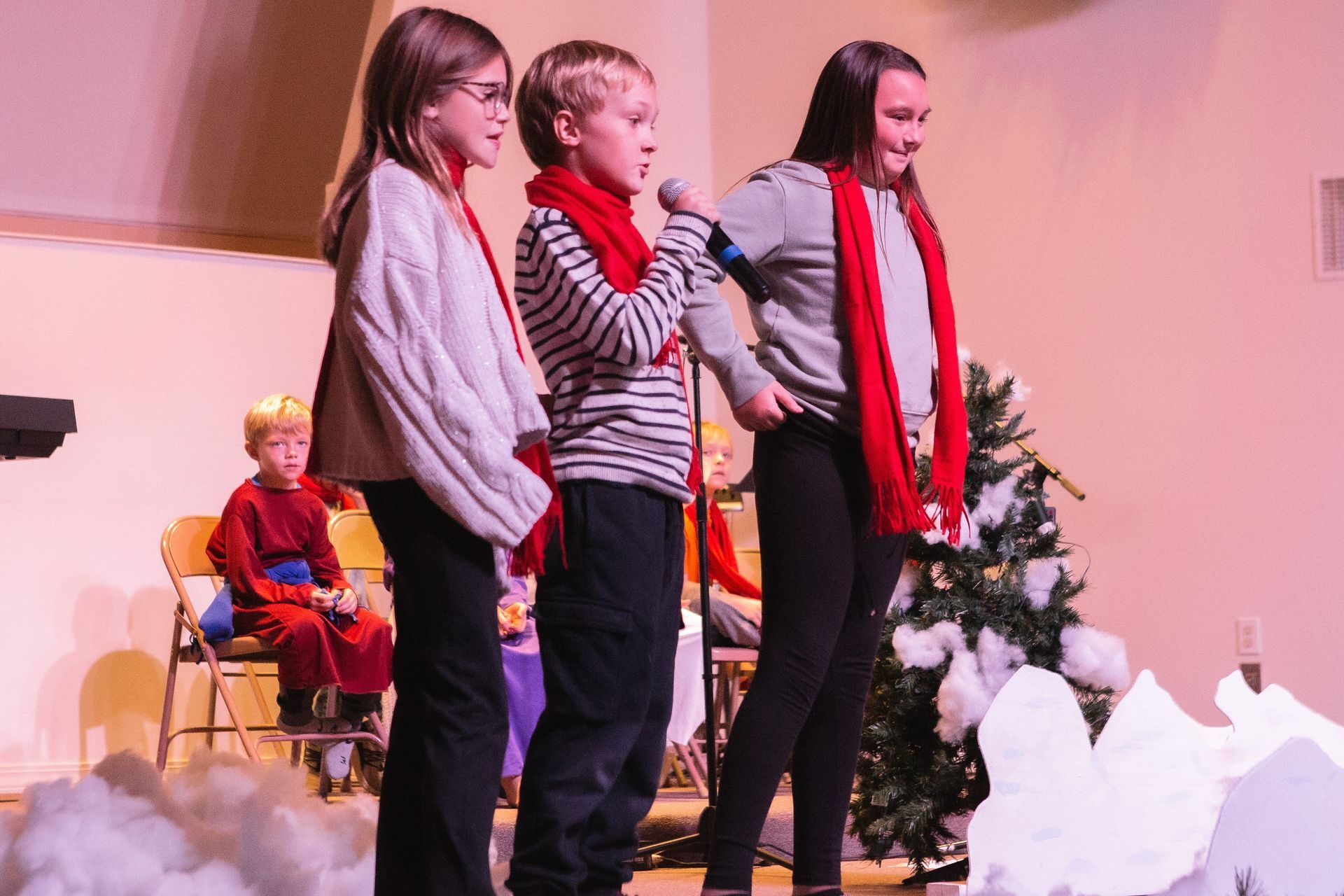 Children singing on stage, wearing scarves. Stage has a Christmas tree and props.