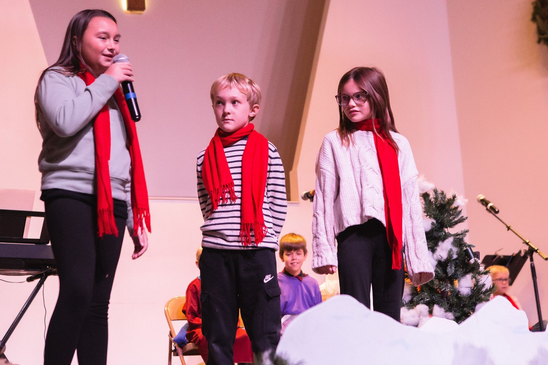 Three children on stage, wearing red scarves. One holds a microphone, speaking. Snowy set.