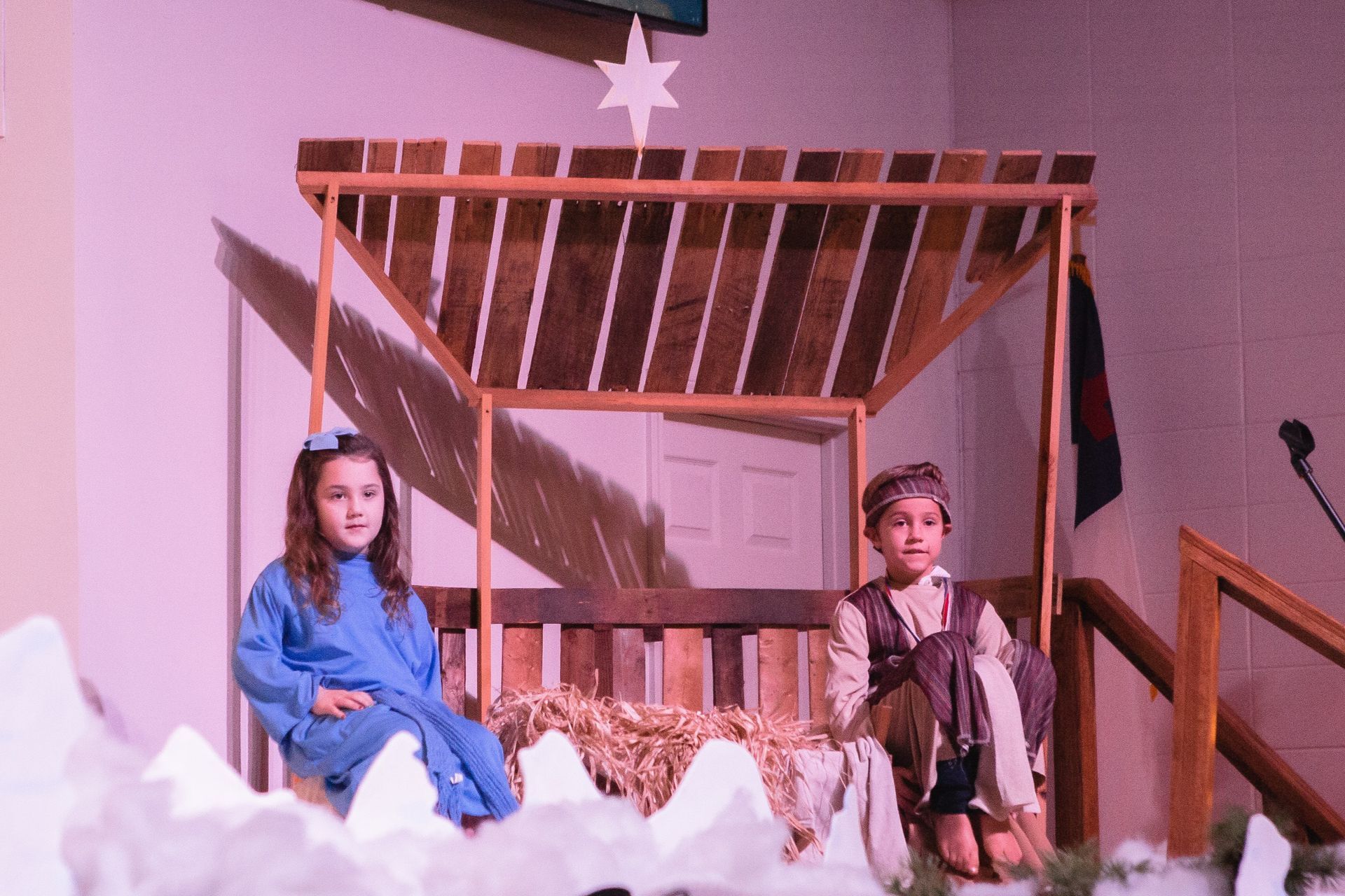Children in costumes sit near a wooden structure on a stage.
