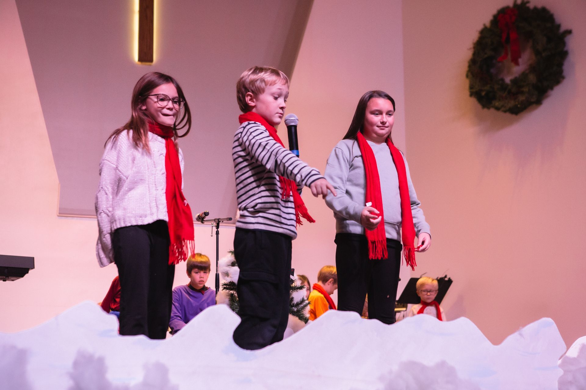 Children singing on a stage with fake snow, wearing red scarves. Wreath on the wall.