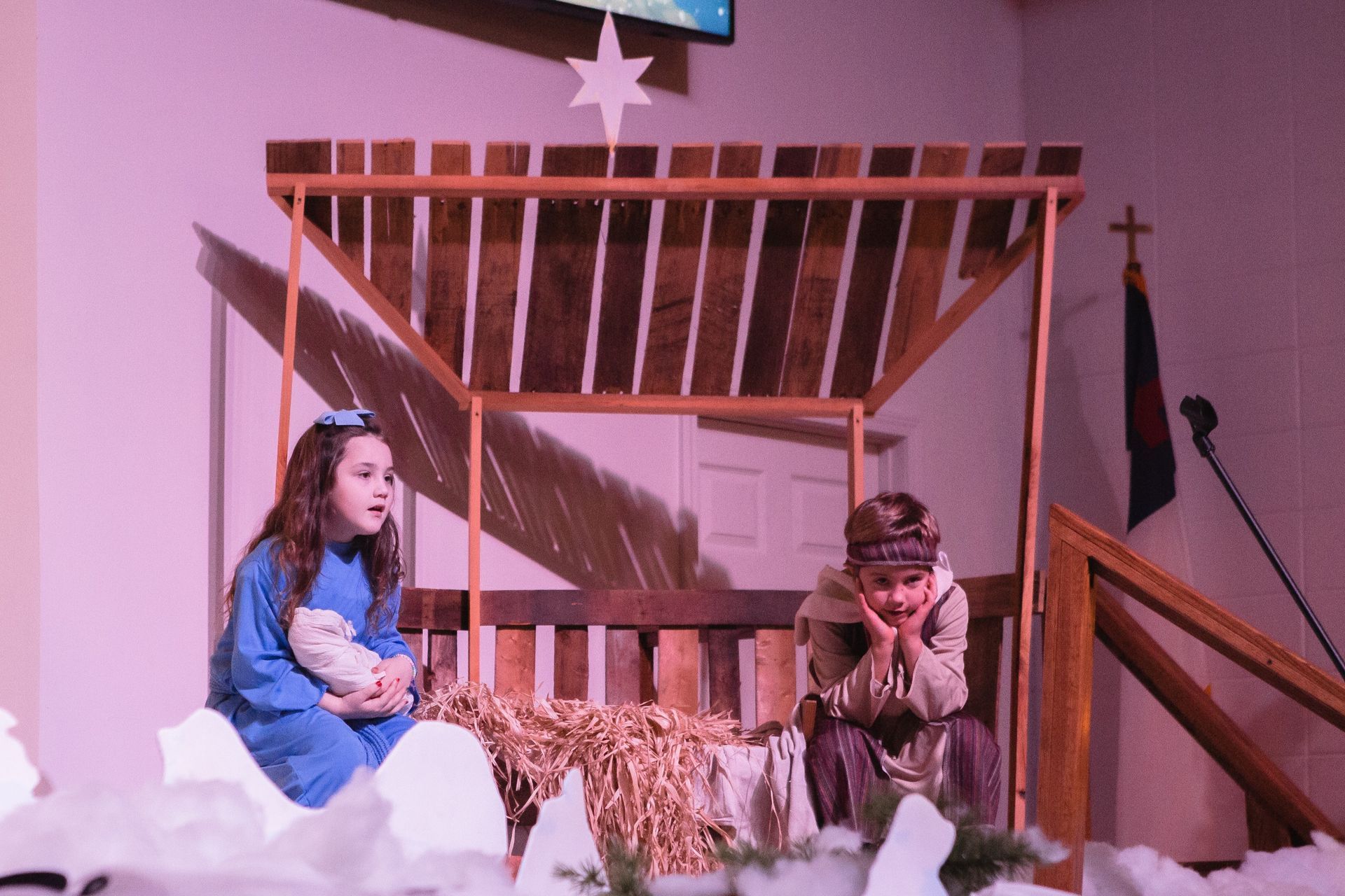 Children in a nativity play scene: girl in blue holding a lamb, boy in brown, manger backdrop.