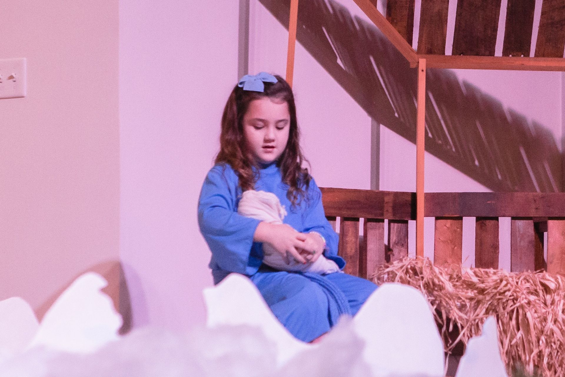 Girl in blue robe holding a stuffed animal, seated in a Nativity scene.