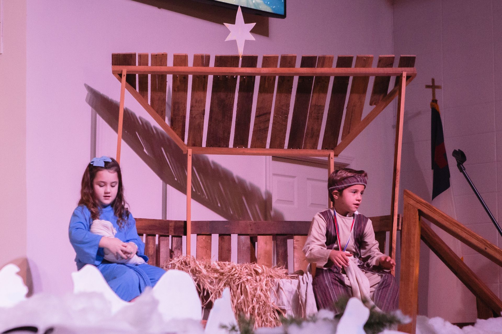Two children in costumes on a stage, acting in a nativity scene; one holds a lamb.