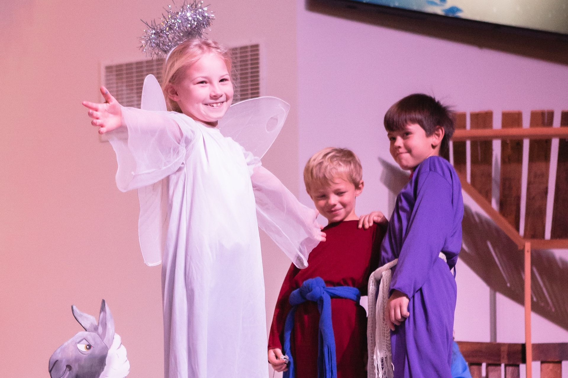 Children dressed in costumes on a stage. One is an angel with a thumbs up, two are in biblical attire.