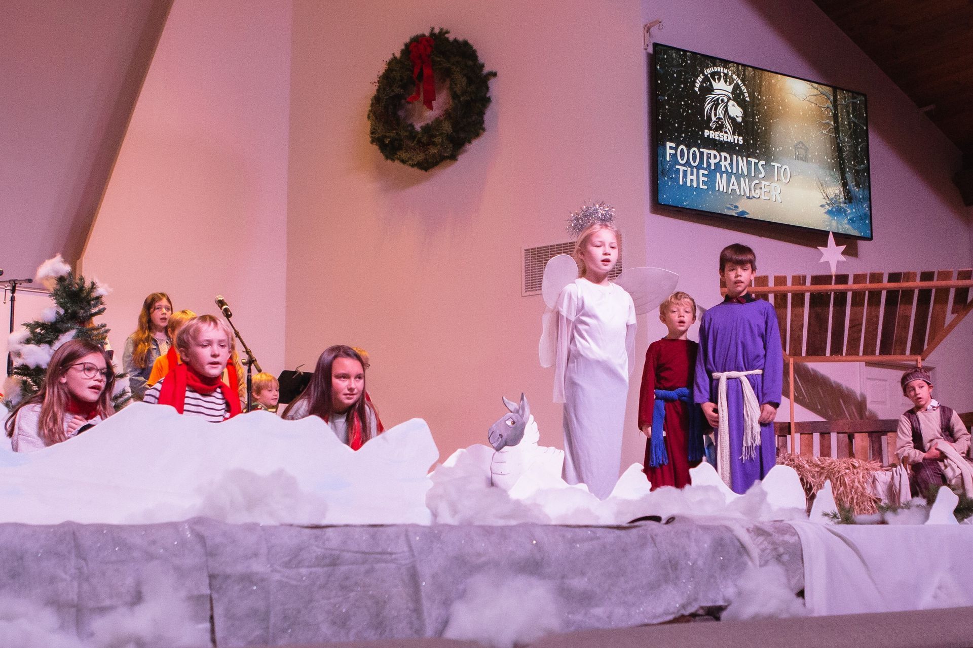 Children performing a nativity play on stage, wearing costumes and standing in front of a screen.