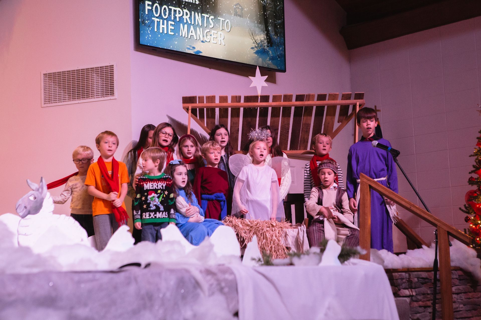 Children performing a Christmas nativity play on stage, with a manger scene and a projected title.