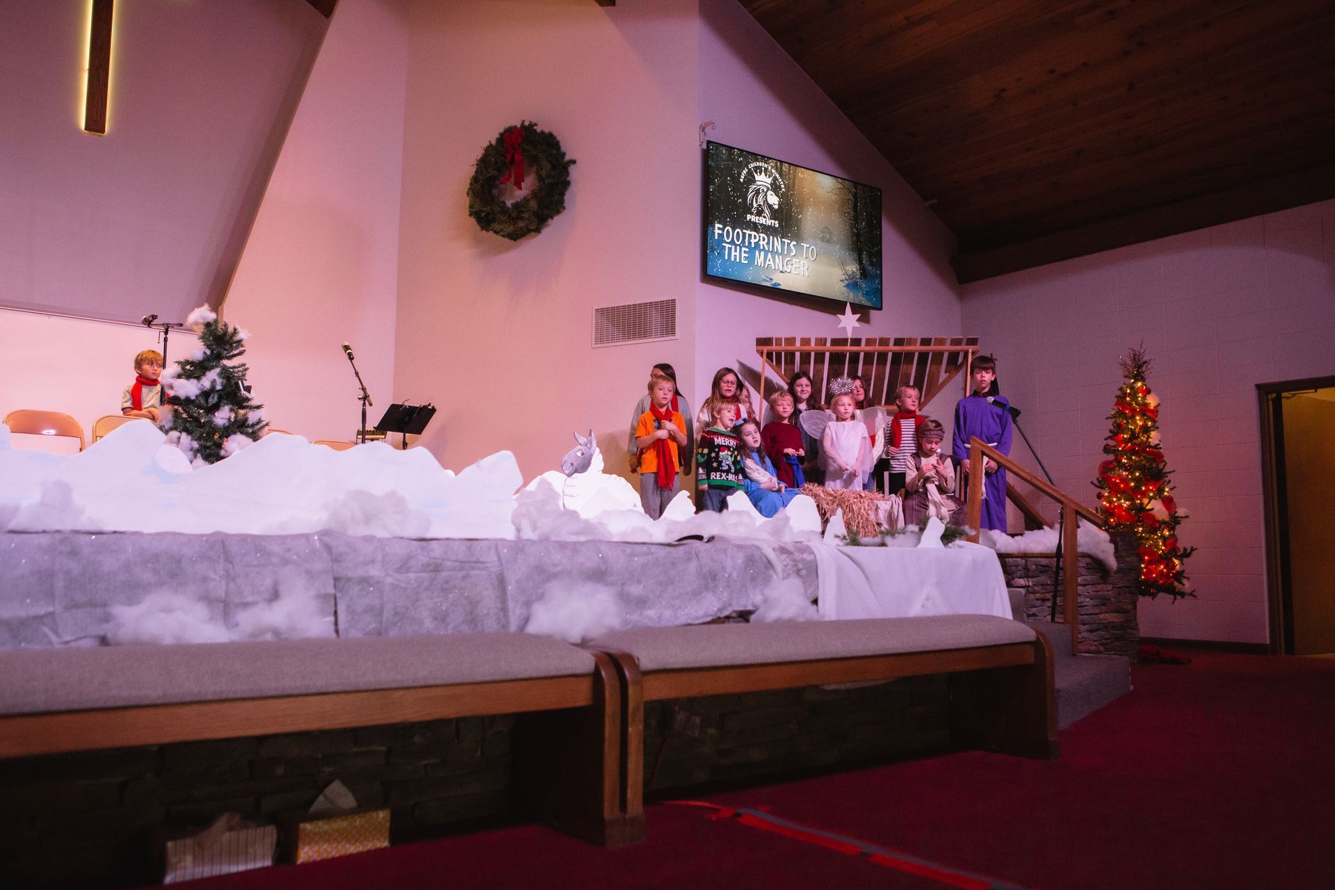 Children performing on stage in a church decorated for Christmas. Snowy backdrop, Christmas trees, and a screen in the background.
