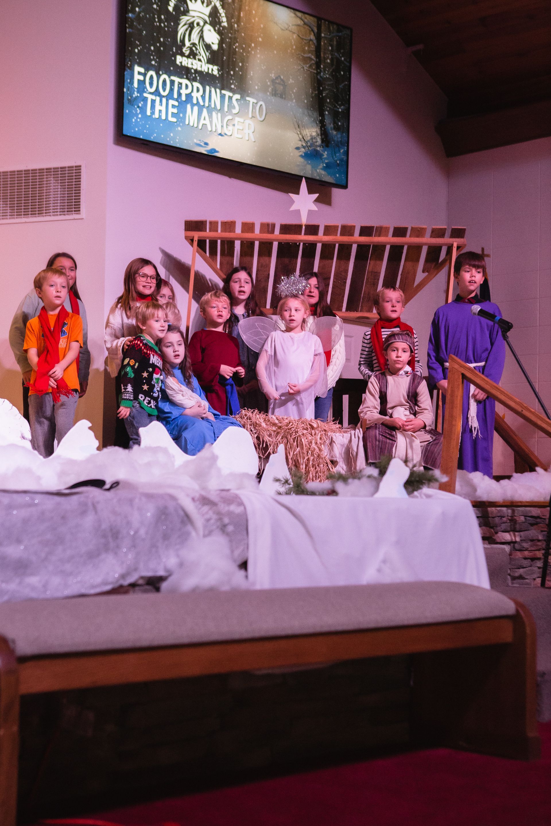 Children in costumes perform a nativity scene on a stage in a church. A screen shows the words 